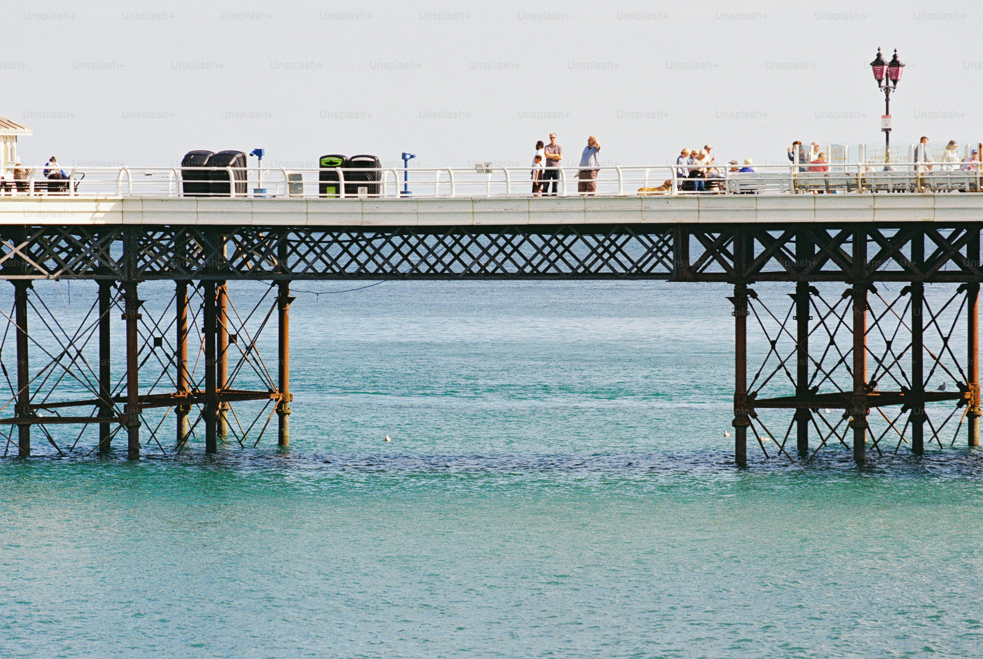 People walking on a pier supported by iron girders above calm blue sea water