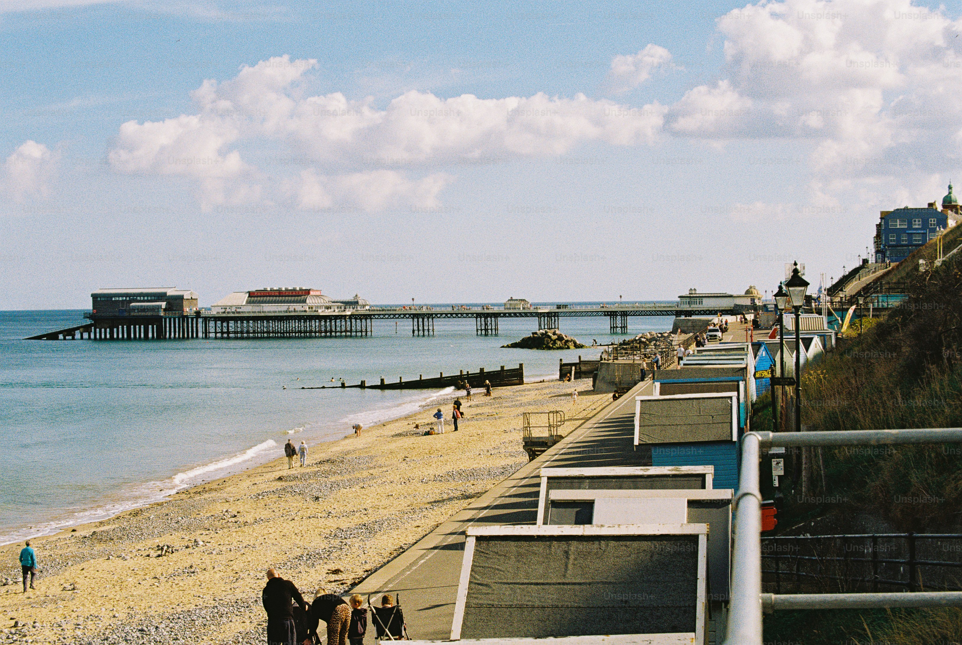 People on a shingle beach and promenade near Cromer Pier under a blue sky