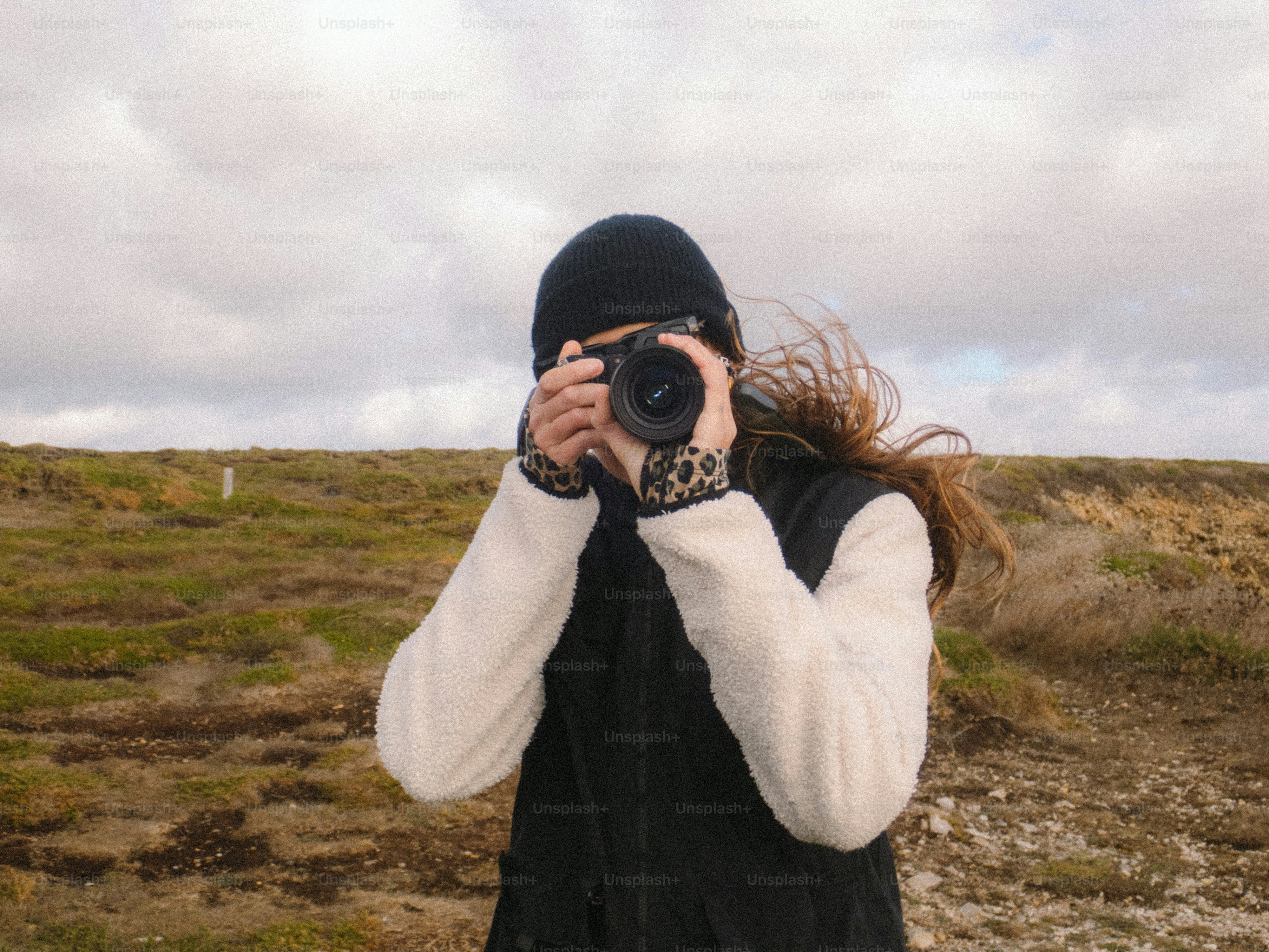 A person in a black beanie and fleece jacket taking a photograph on a grassy hillside
