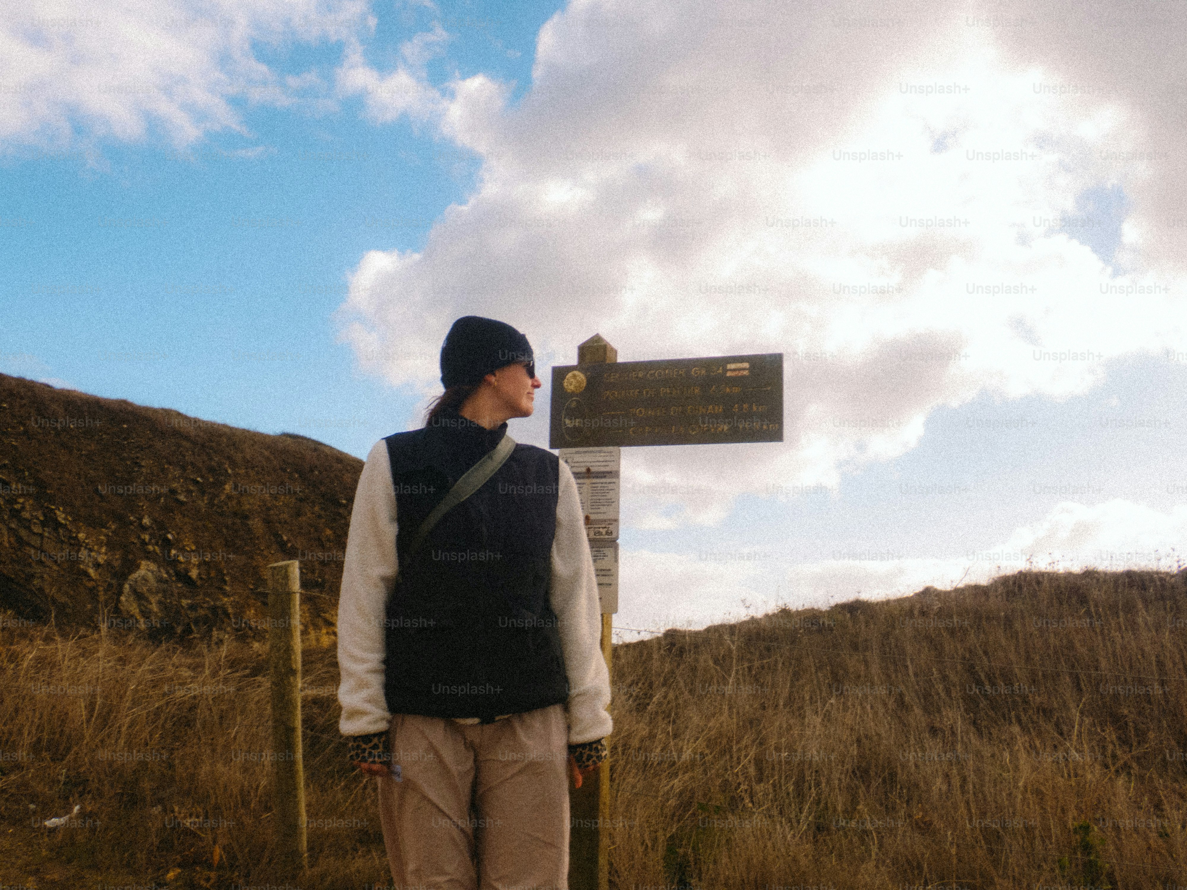 A person in a beanie and vest standing by a trail sign on a grassy hill