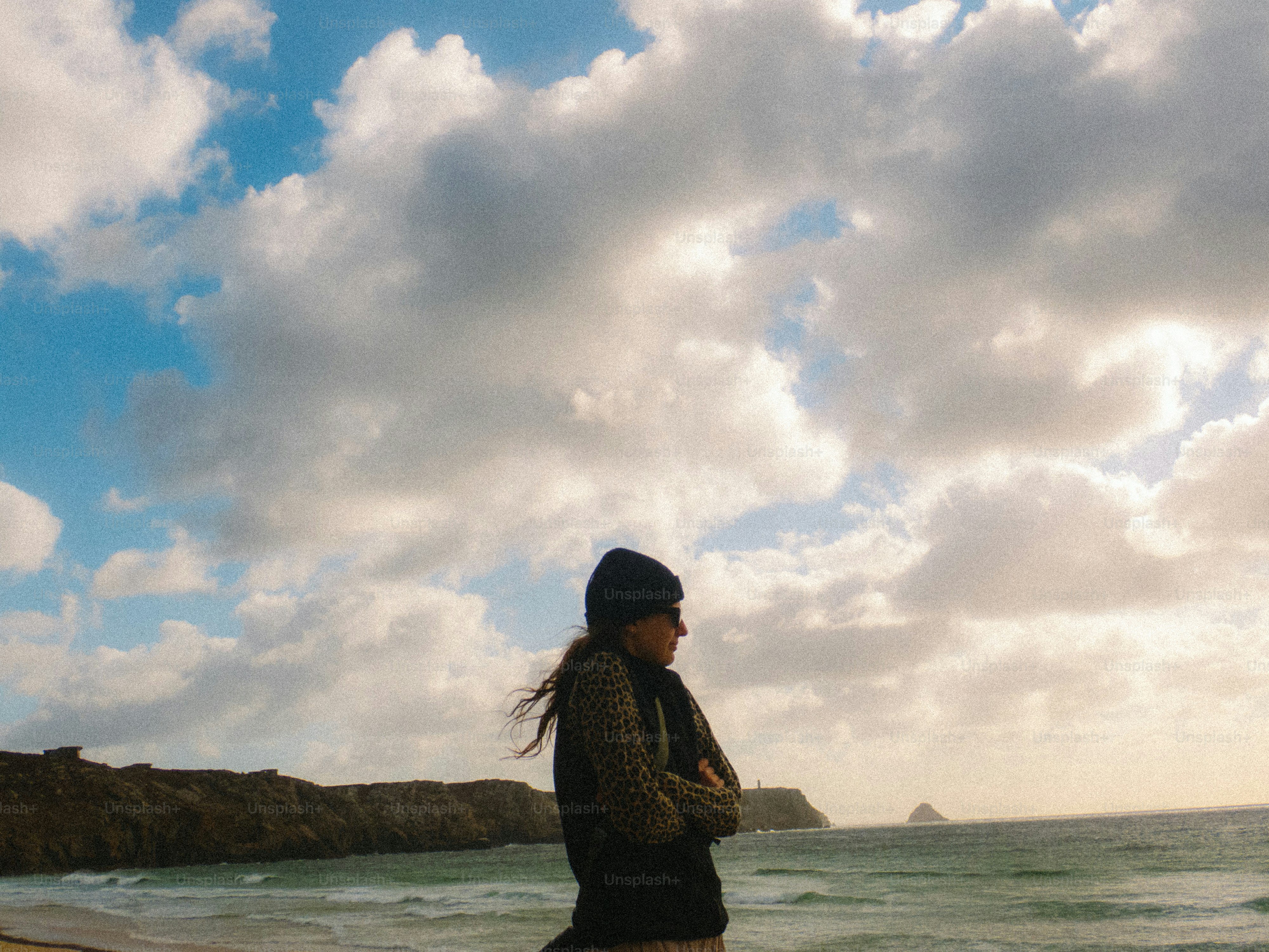 A person in a beanie and leopard print top on a beach under a cloudy sky