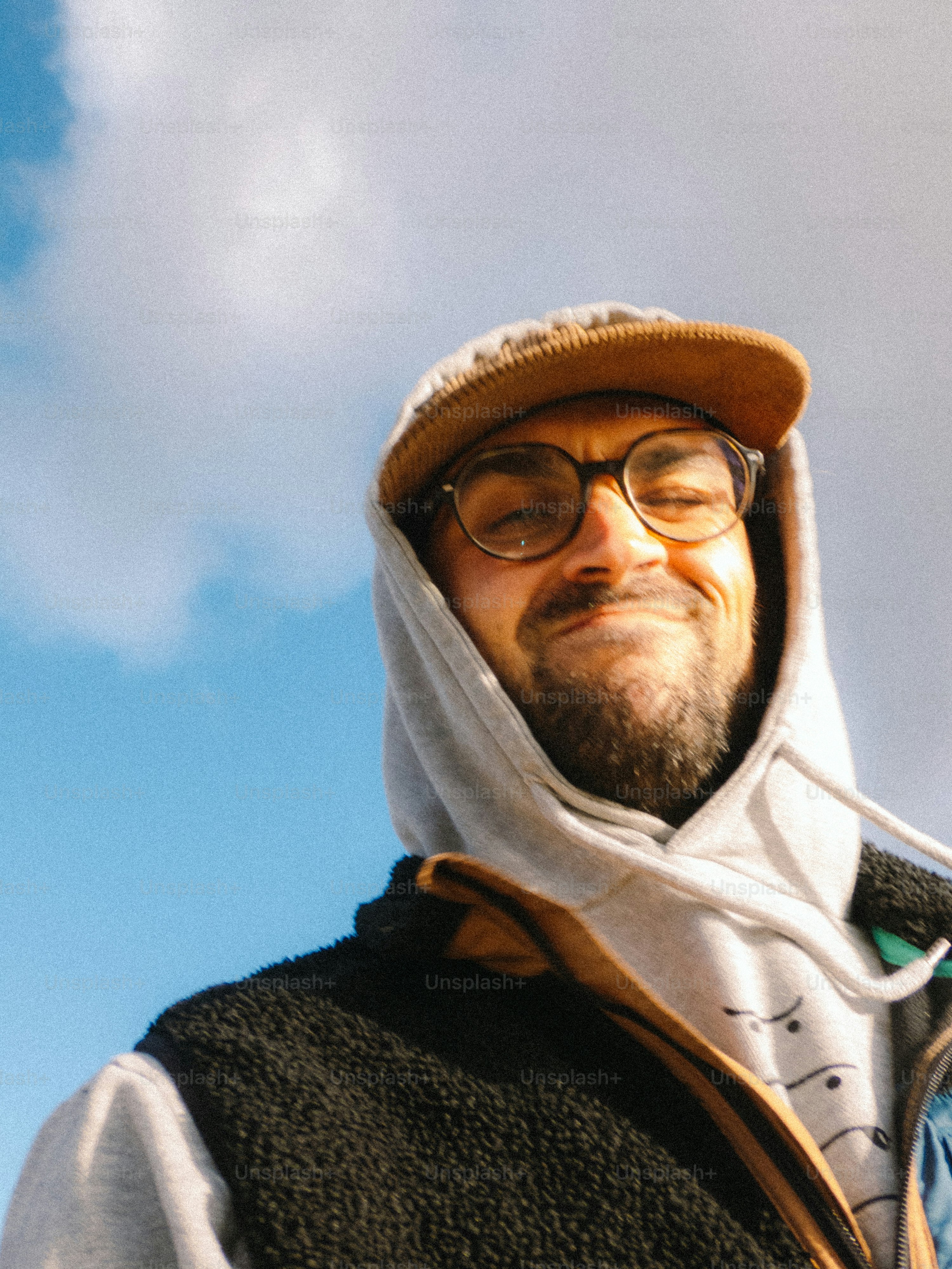 A man with a beard wearing a grey hoodie and brown cap against a blue sky
