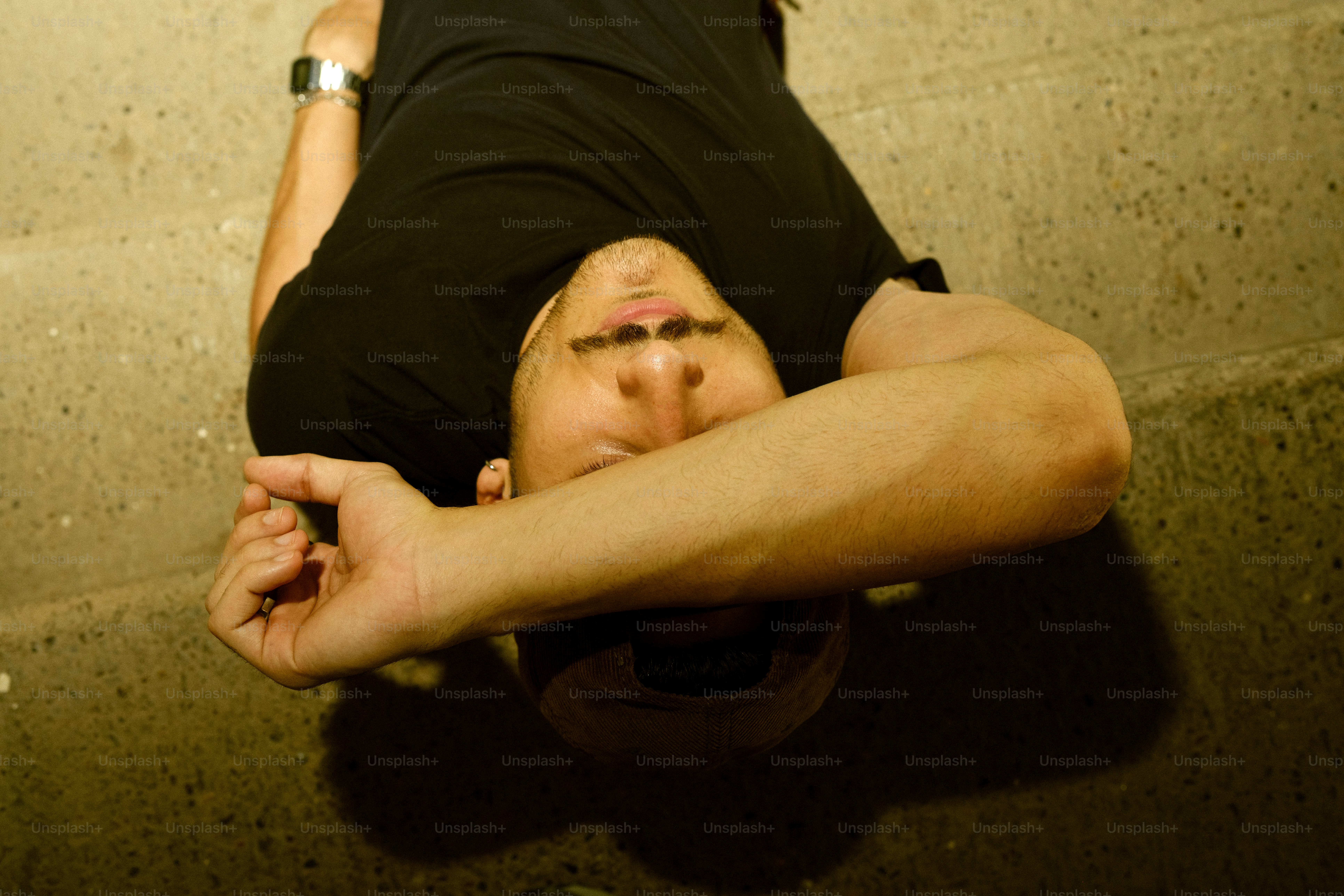 Eine Person in einem schwarzen T-Shirt, die auf einem Betonboden liegt, mit einem Arm, der ihr Gesicht bedeckt, liegt