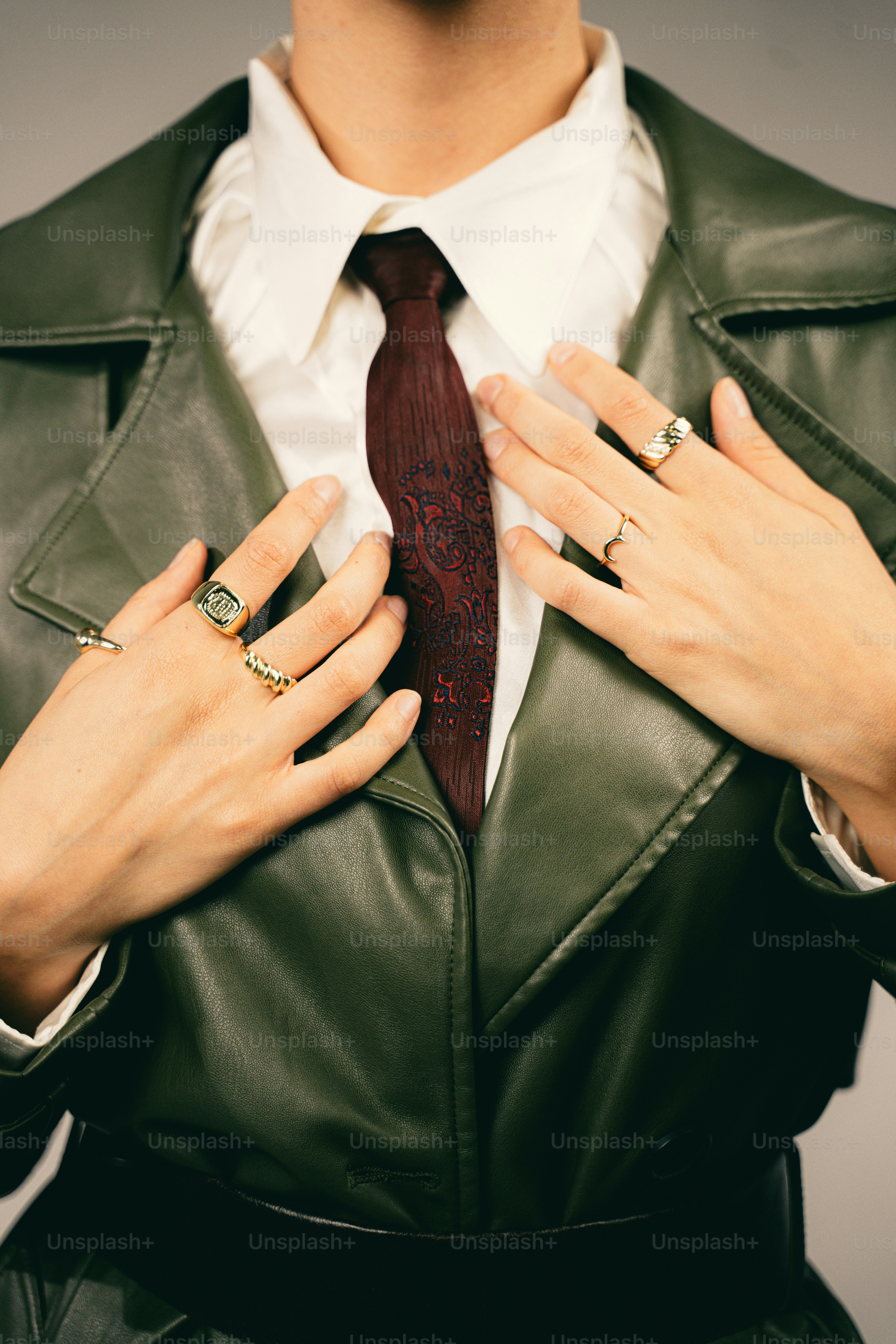 A person wearing a white shirt, dark patterned tie, and olive green leather jacket with gold rings