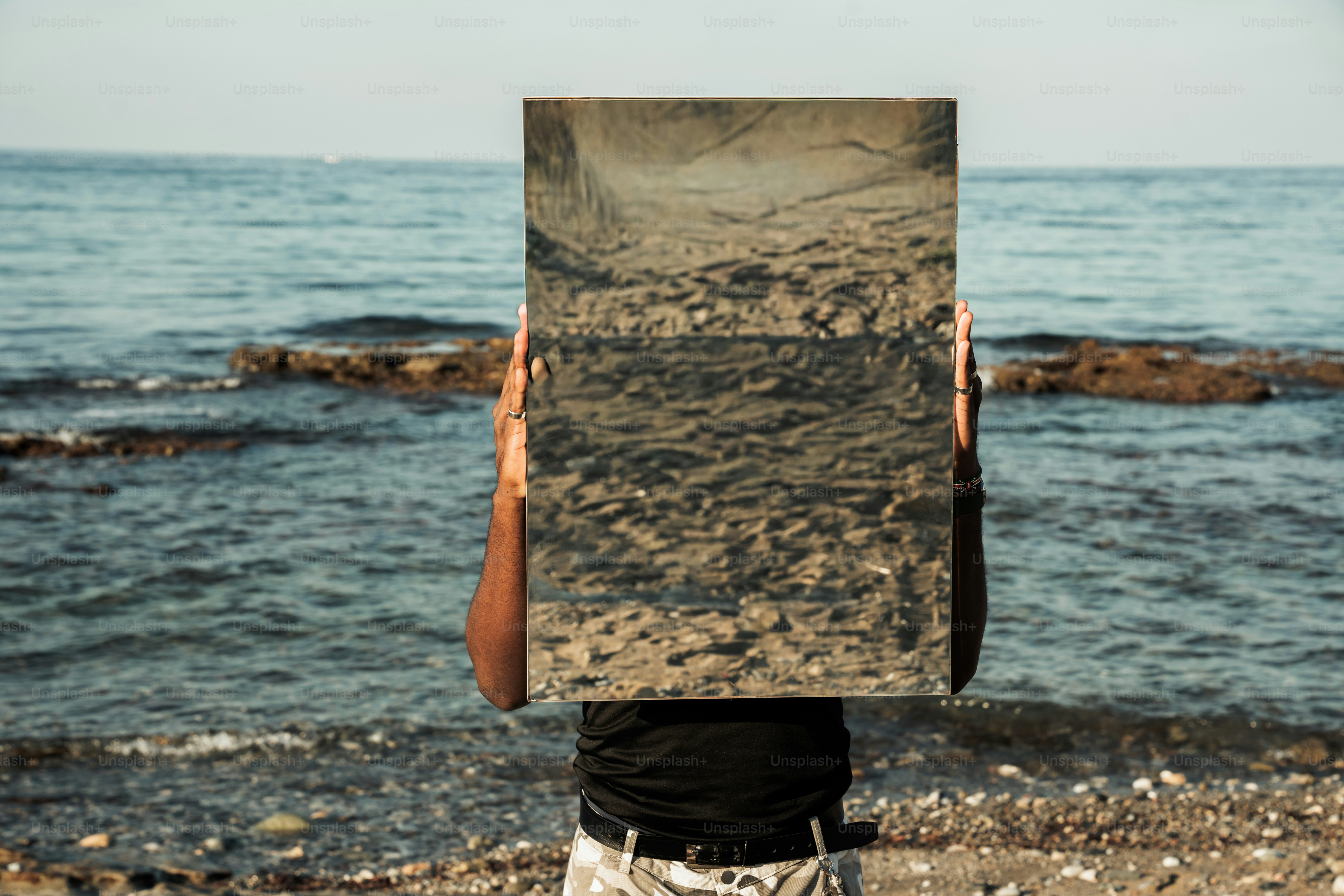 A person holding a large rectangular mirror reflecting the rocky beach and sea