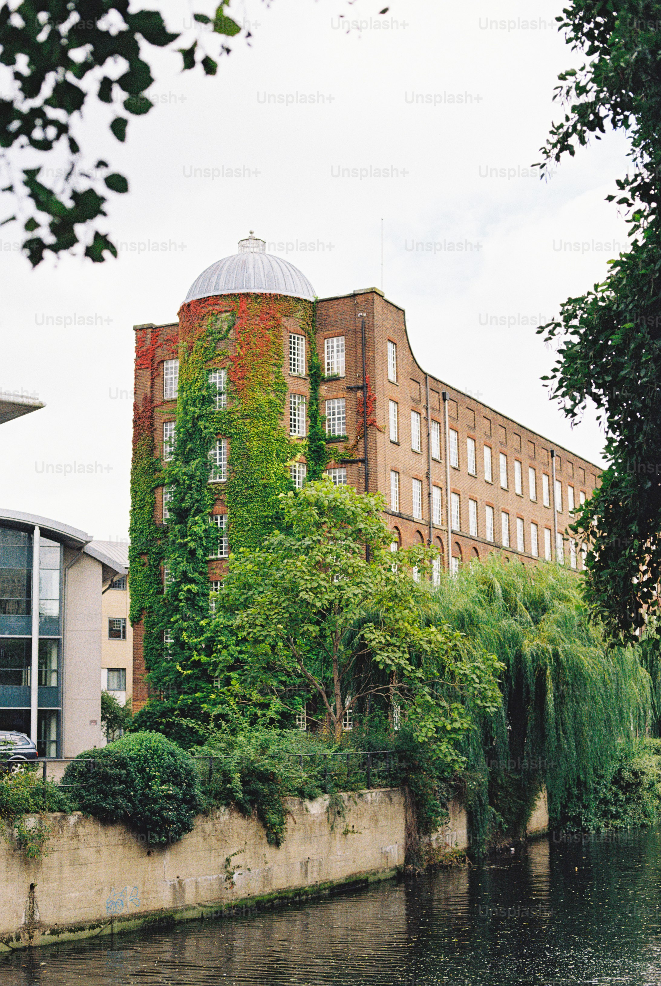 A brick building with a domed tower covered in green and red ivy beside a canal