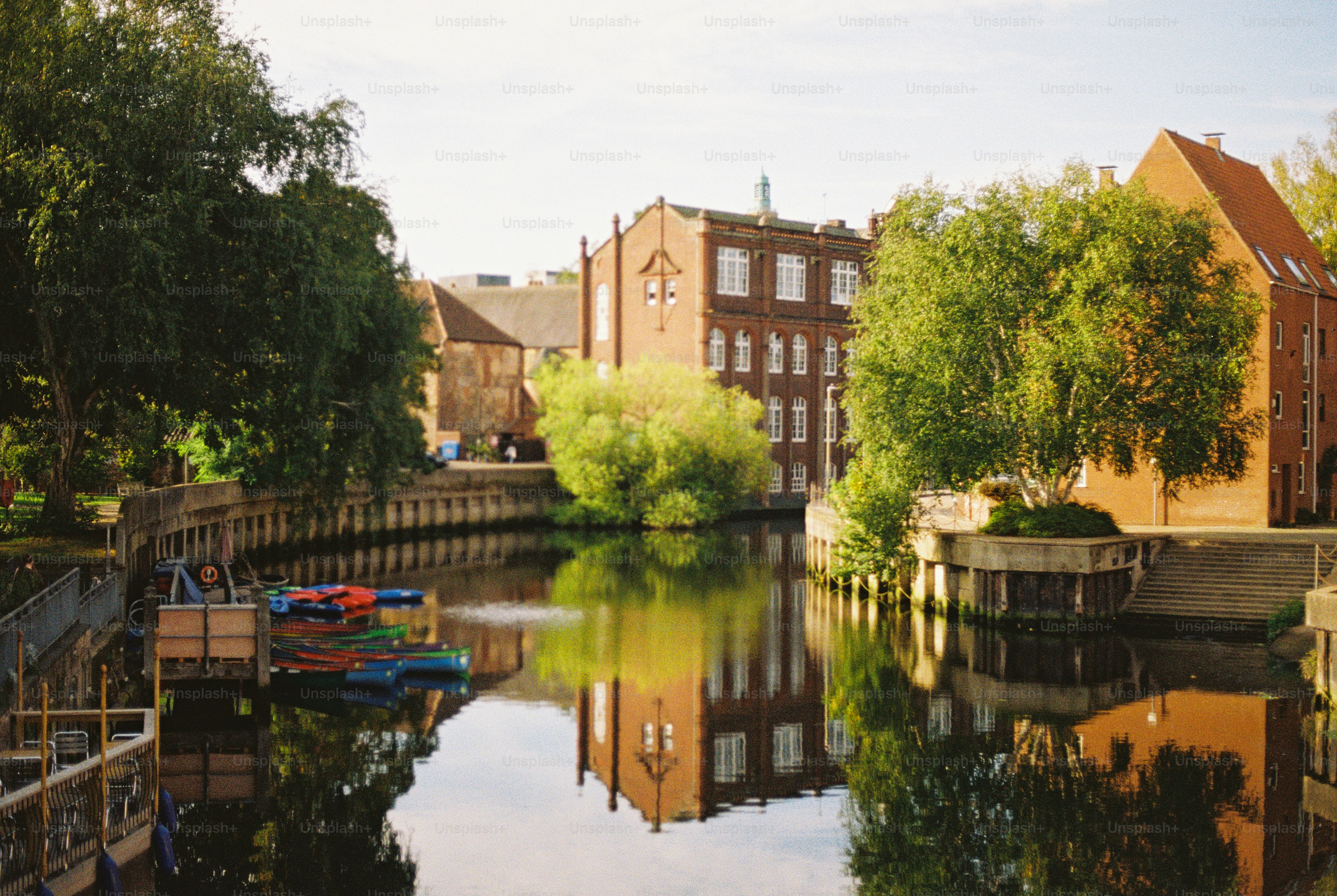 A calm river reflecting a red brick building and green trees on a sunny day