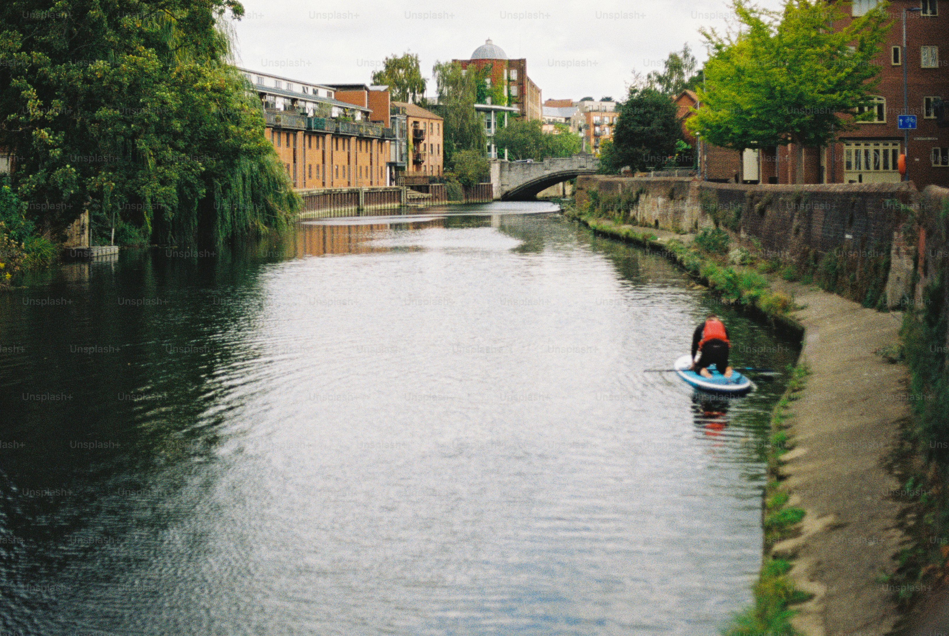 A person kneeling on a paddleboard on a calm river lined with brick buildings and trees