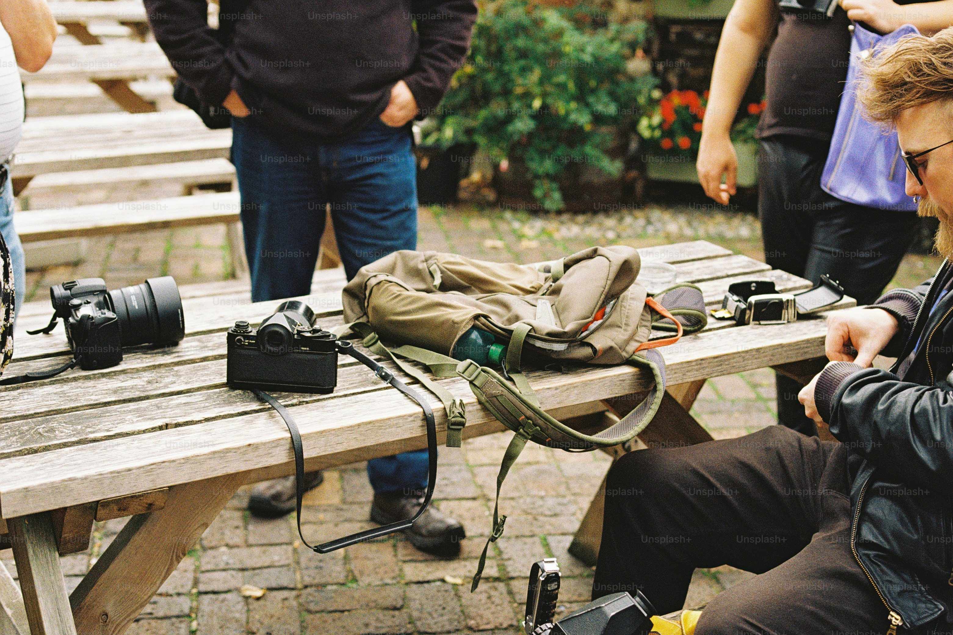 Cameras and a backpack on a wooden picnic table with people standing nearby
