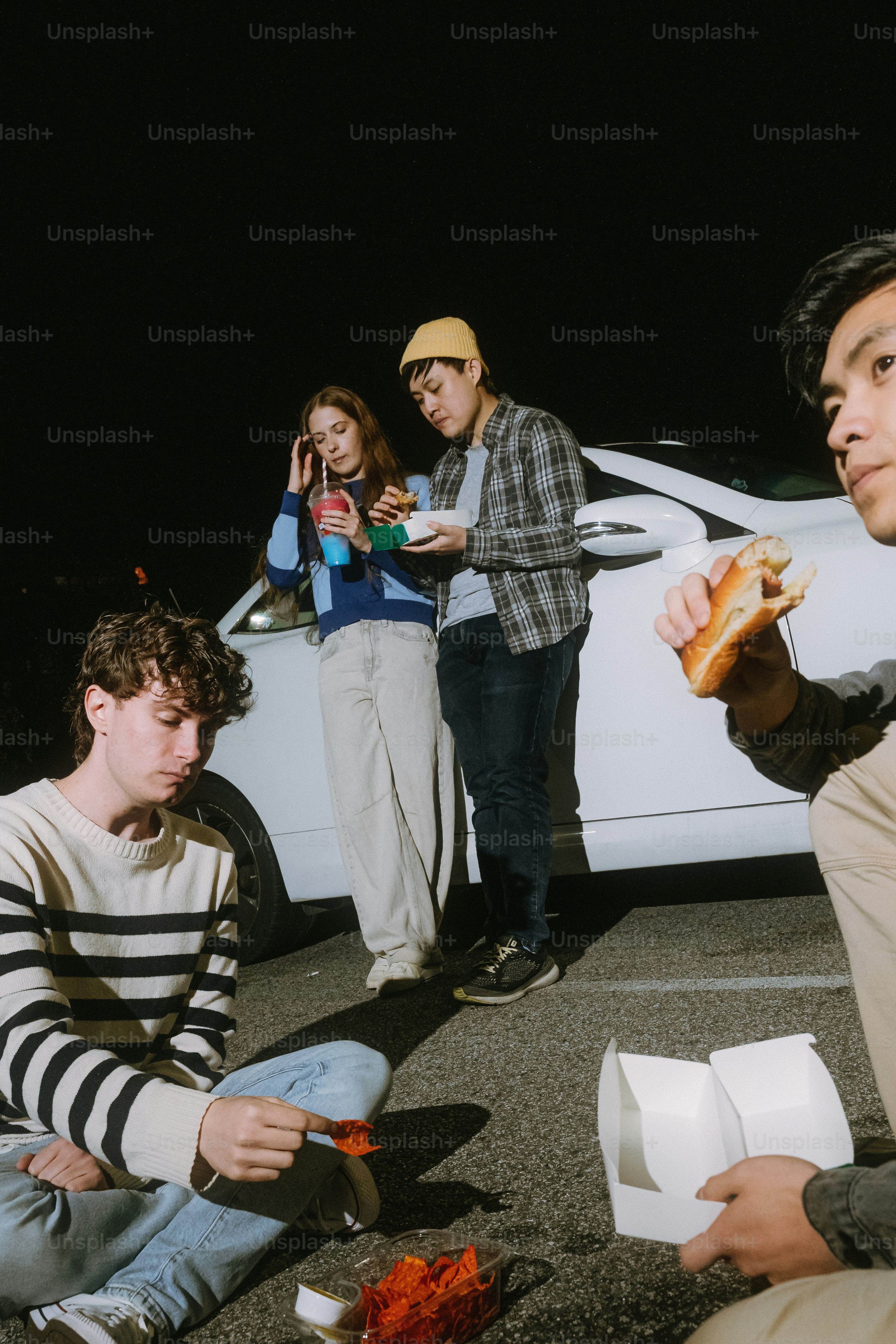 Four people eating fast food in a parking lot at night beside a white car