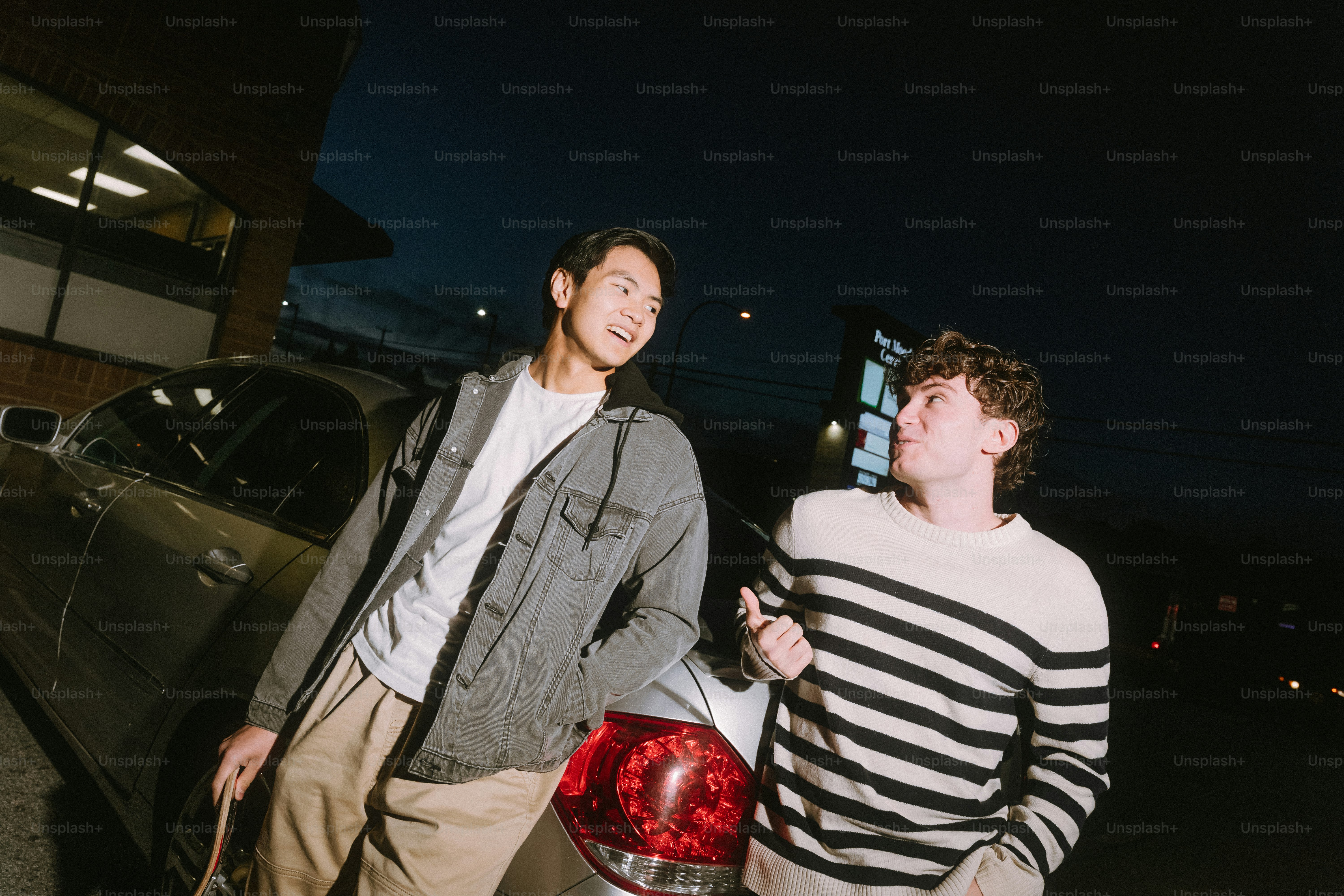 Two young men talking beside a silver car in a parking lot at night