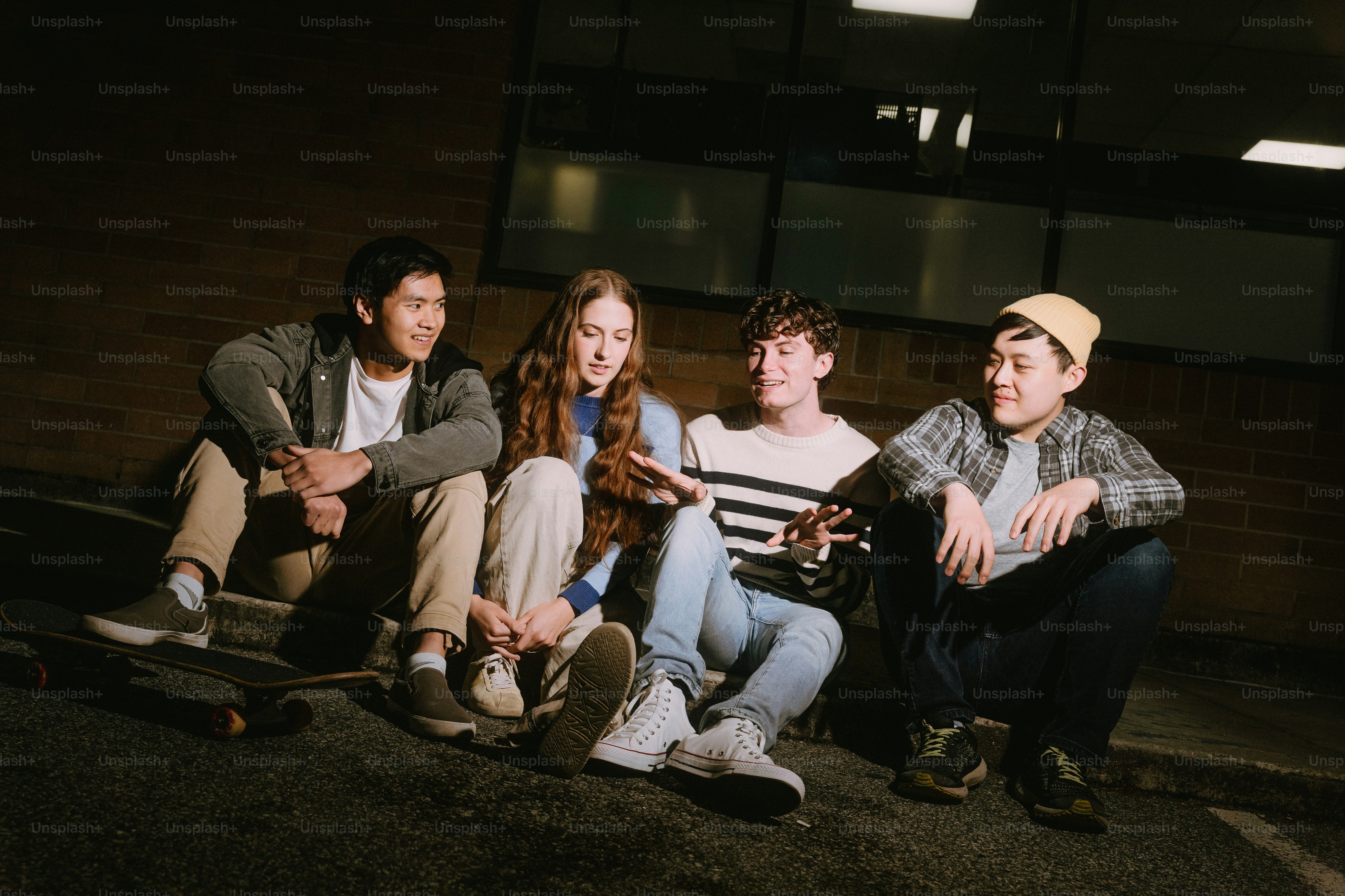 Four young adults sitting on the ground at night near a brick wall with a skateboard