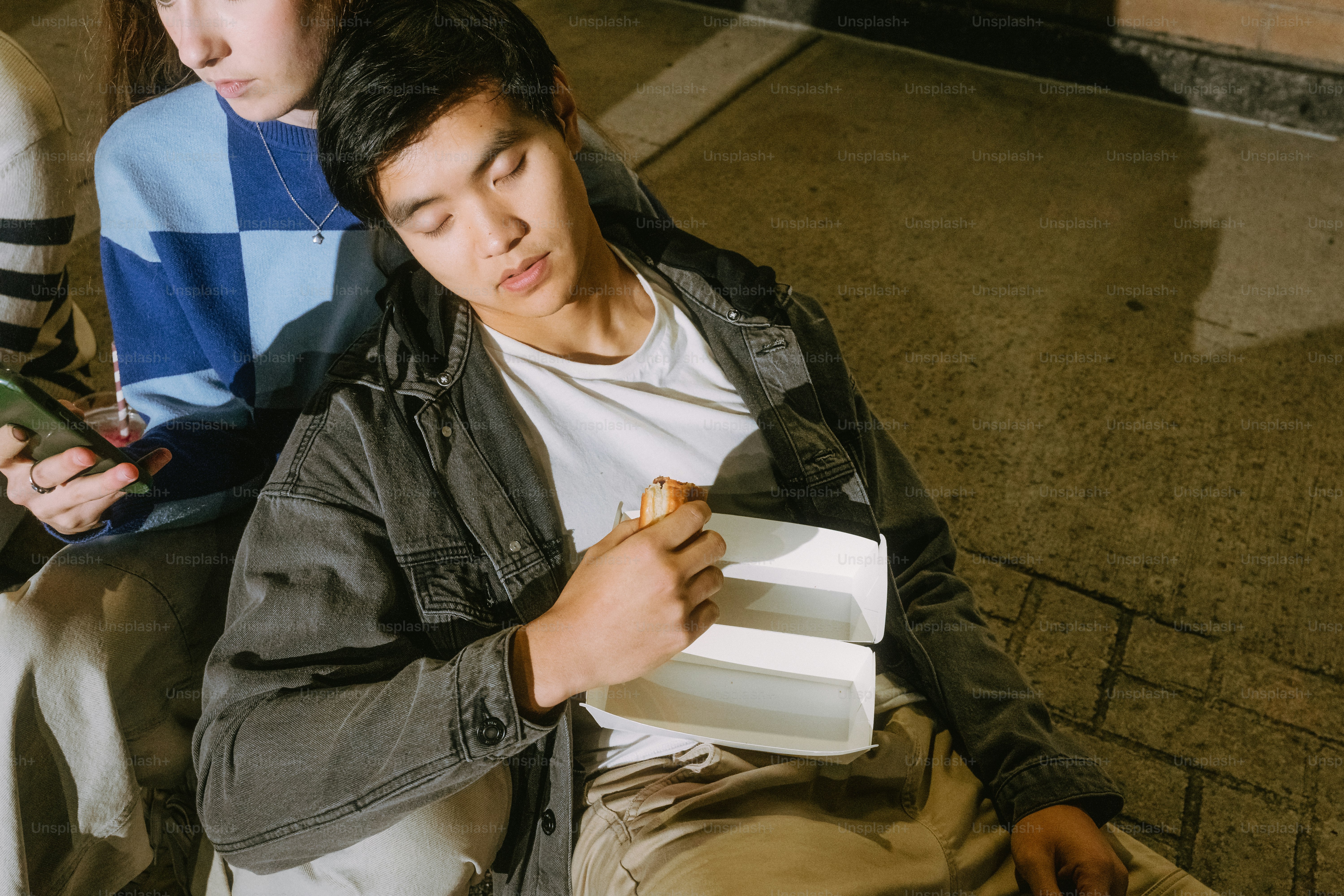 A person eating a snack from a white container while resting against another person on a sidewalk