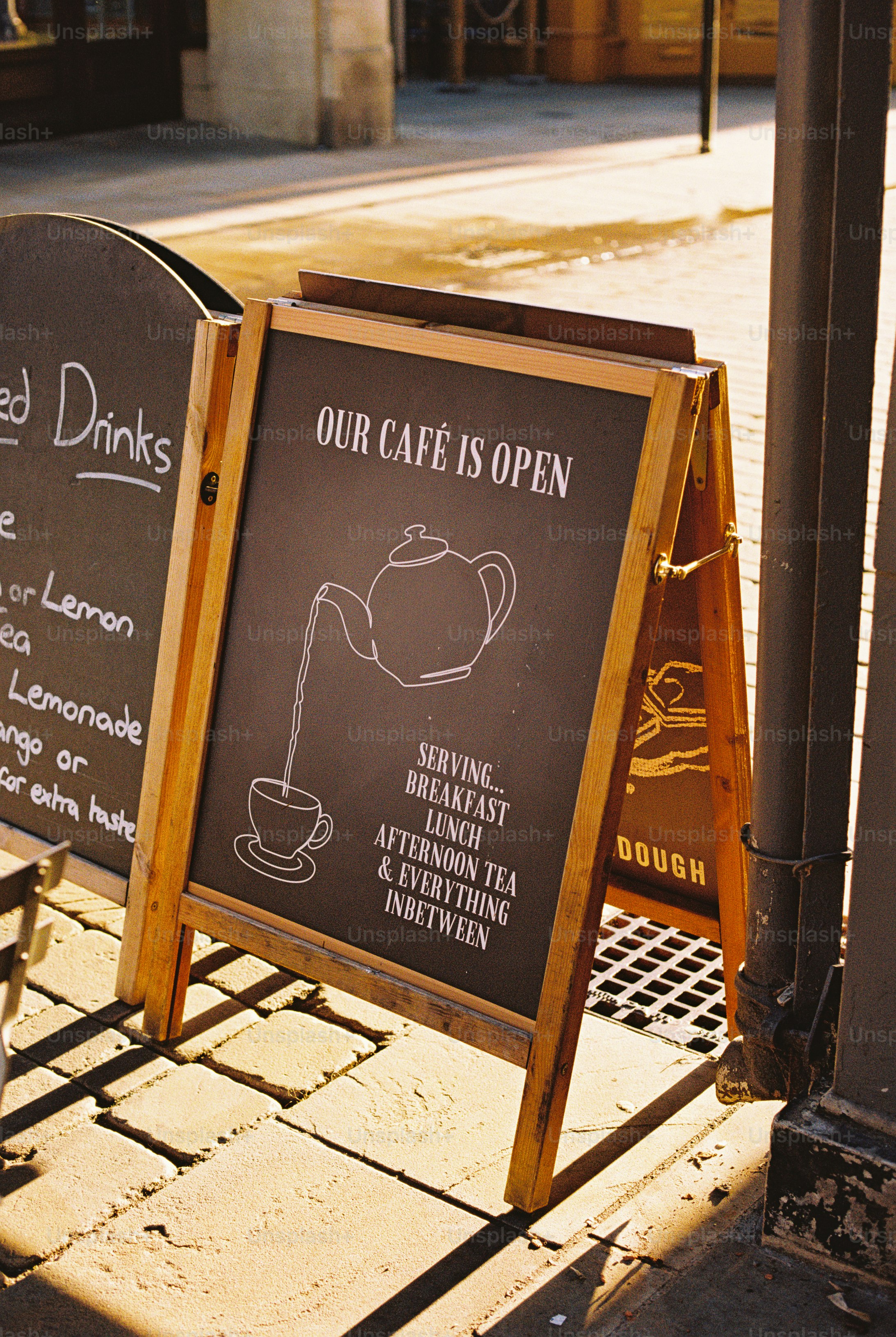 A wooden A-frame chalkboard sign on a sidewalk reading Our Café Is Open with a teapot illustration