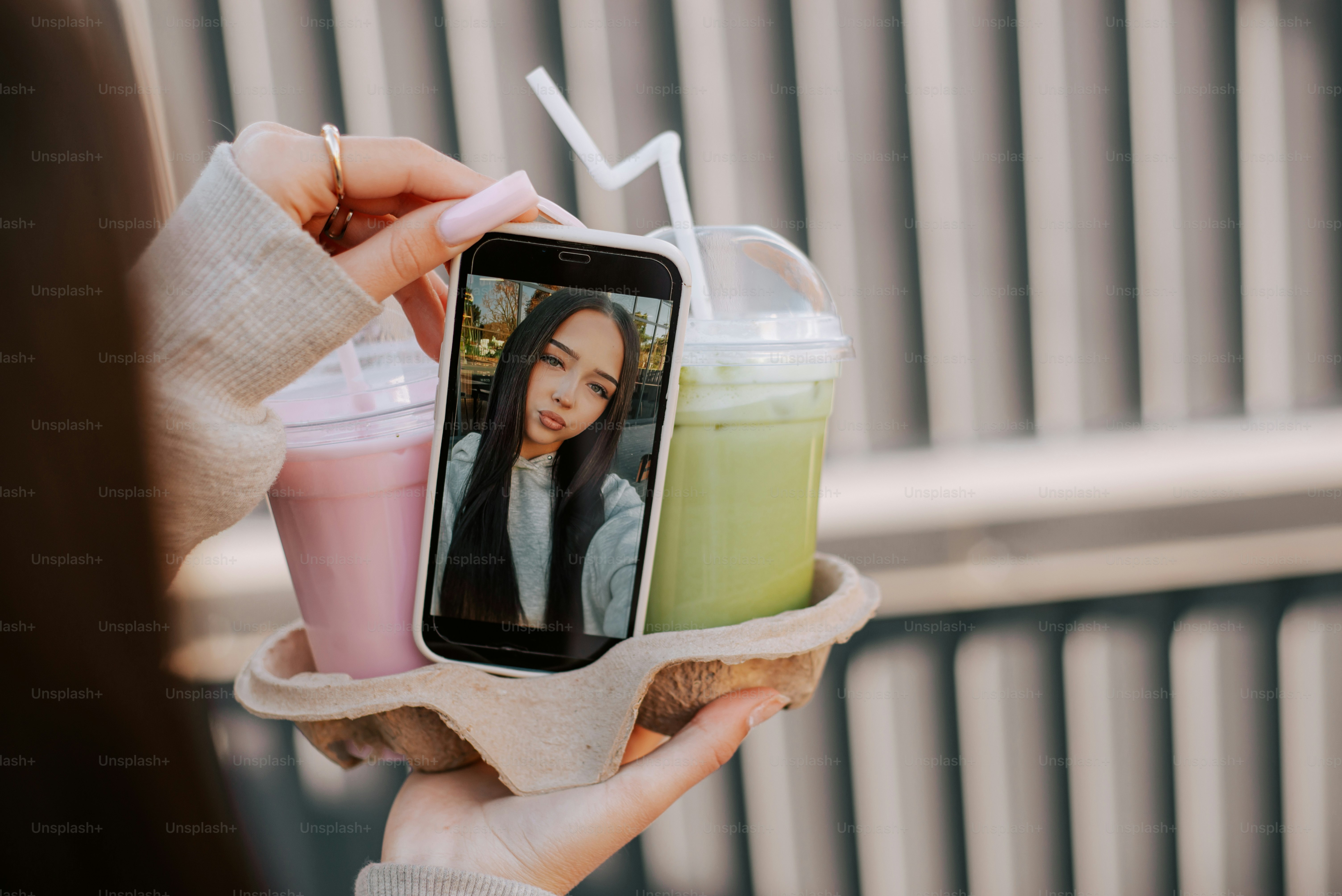 A person holding a cardboard drink carrier with two iced beverages and a smartphone displaying a selfie