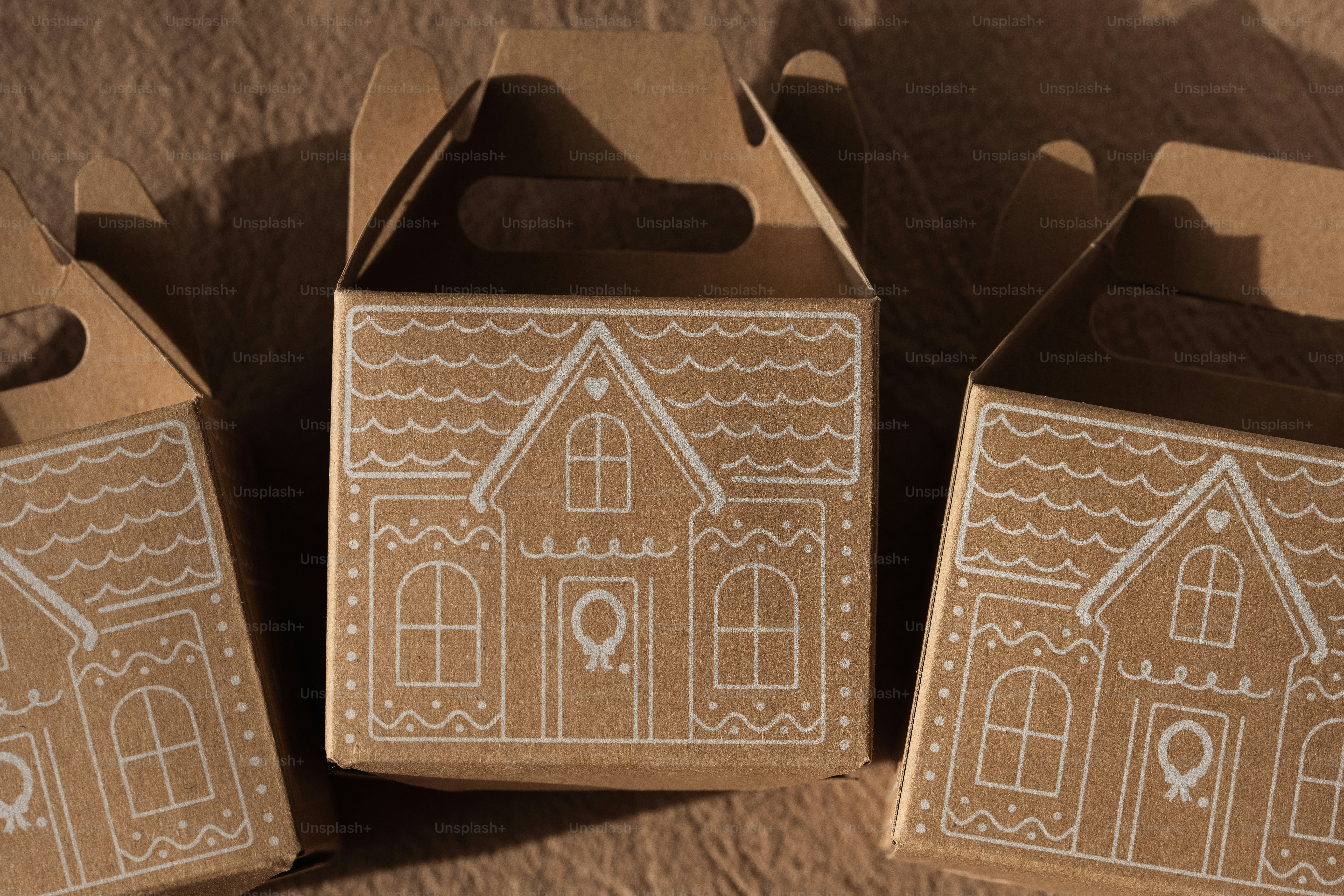 Three brown cardboard gift boxes with white gingerbread house illustrations on a textured surface