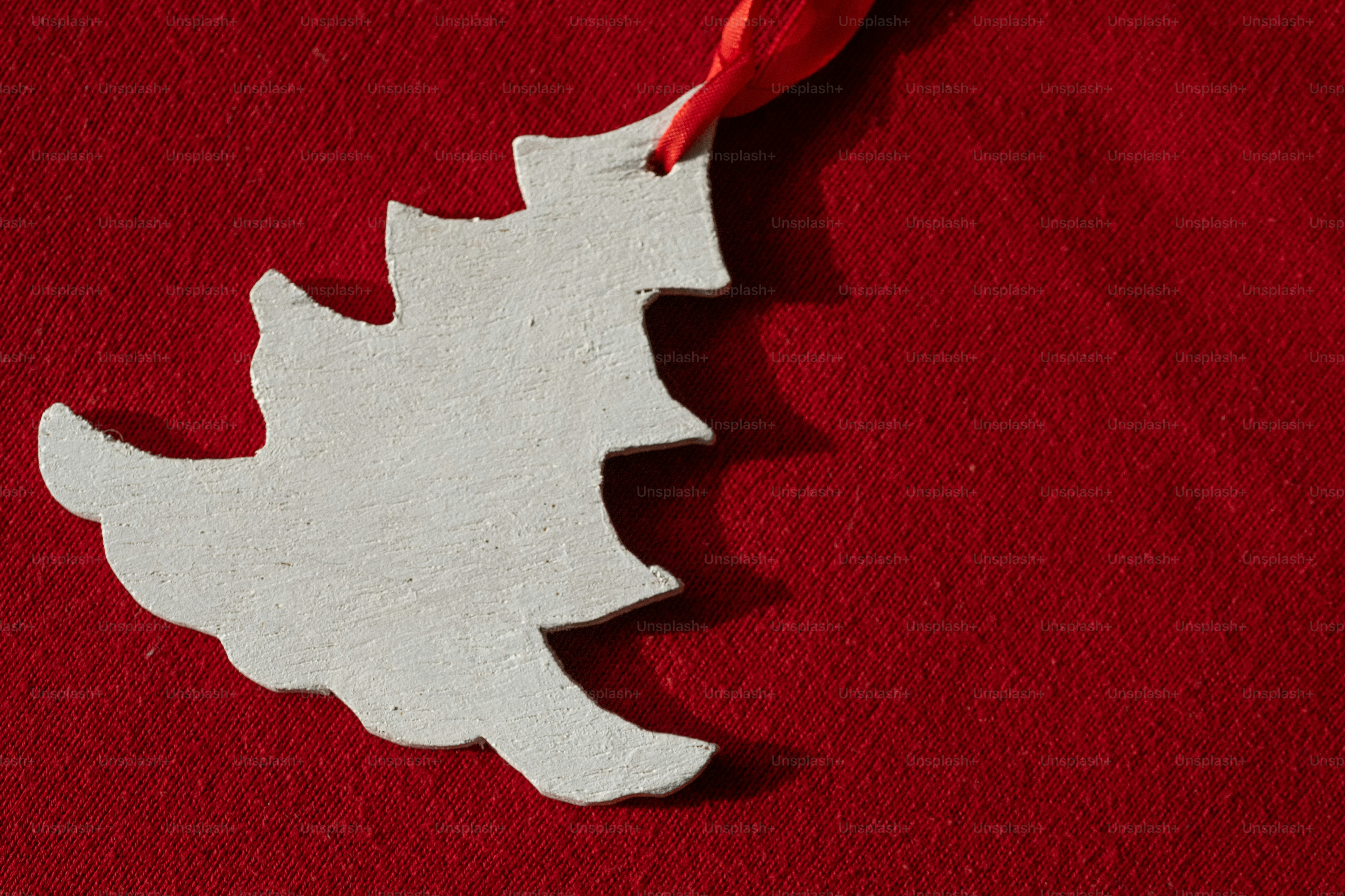 A white wooden Christmas tree ornament hanging by a red ribbon against a red fabric background