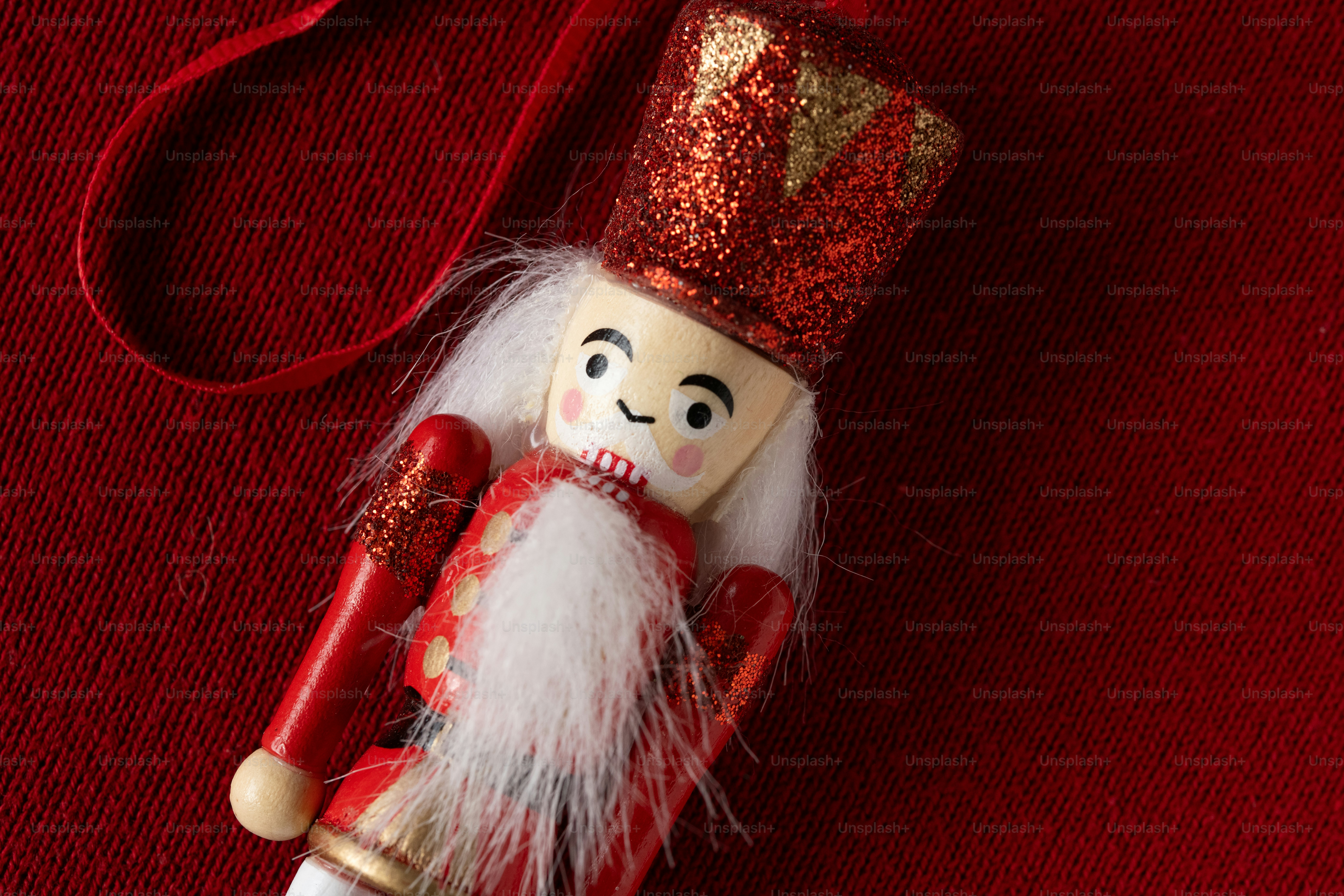 A small wooden nutcracker ornament with a glittery red hat on a textured red fabric background
