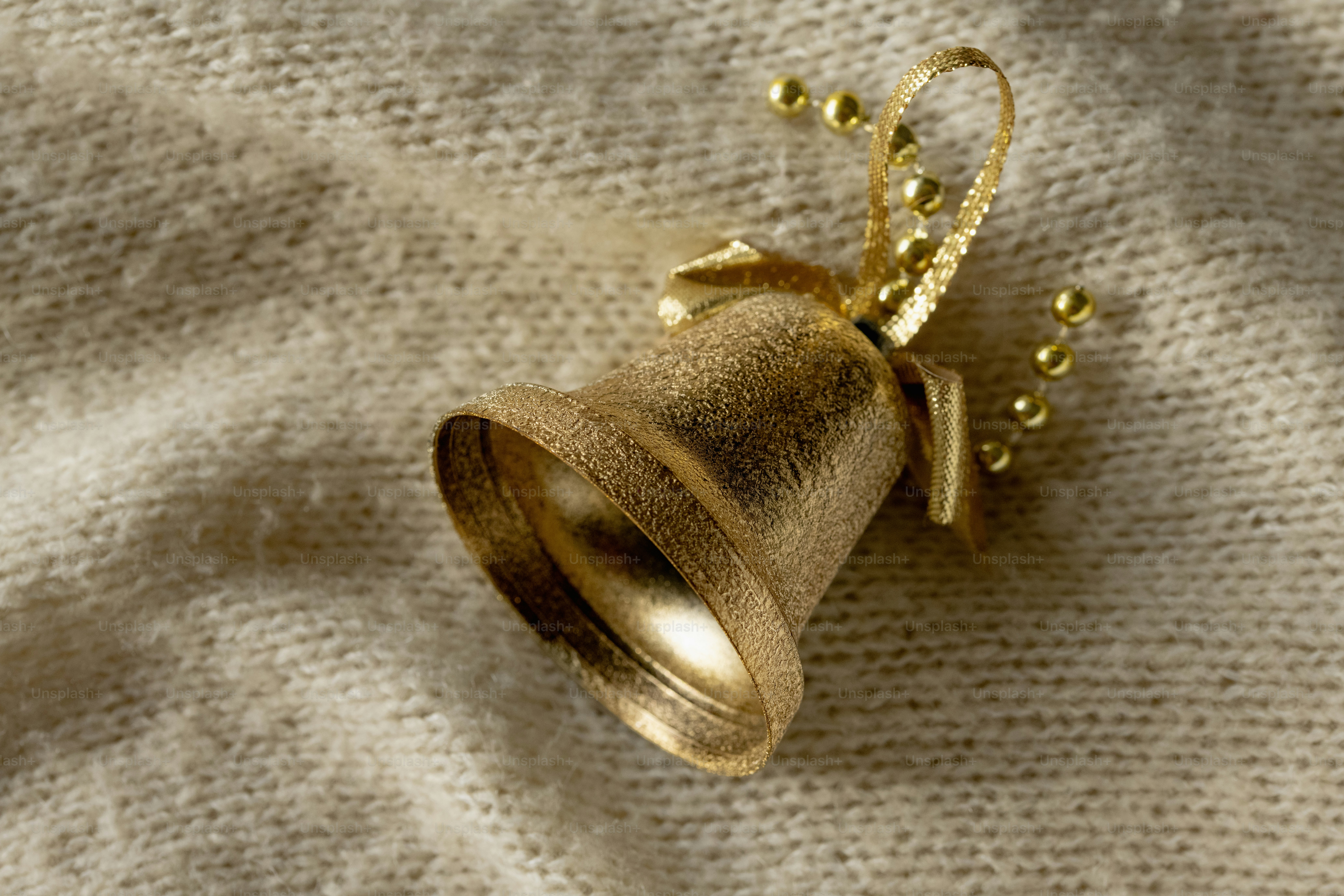 A gold glittery Christmas bell ornament with a beaded string on a beige knitted fabric