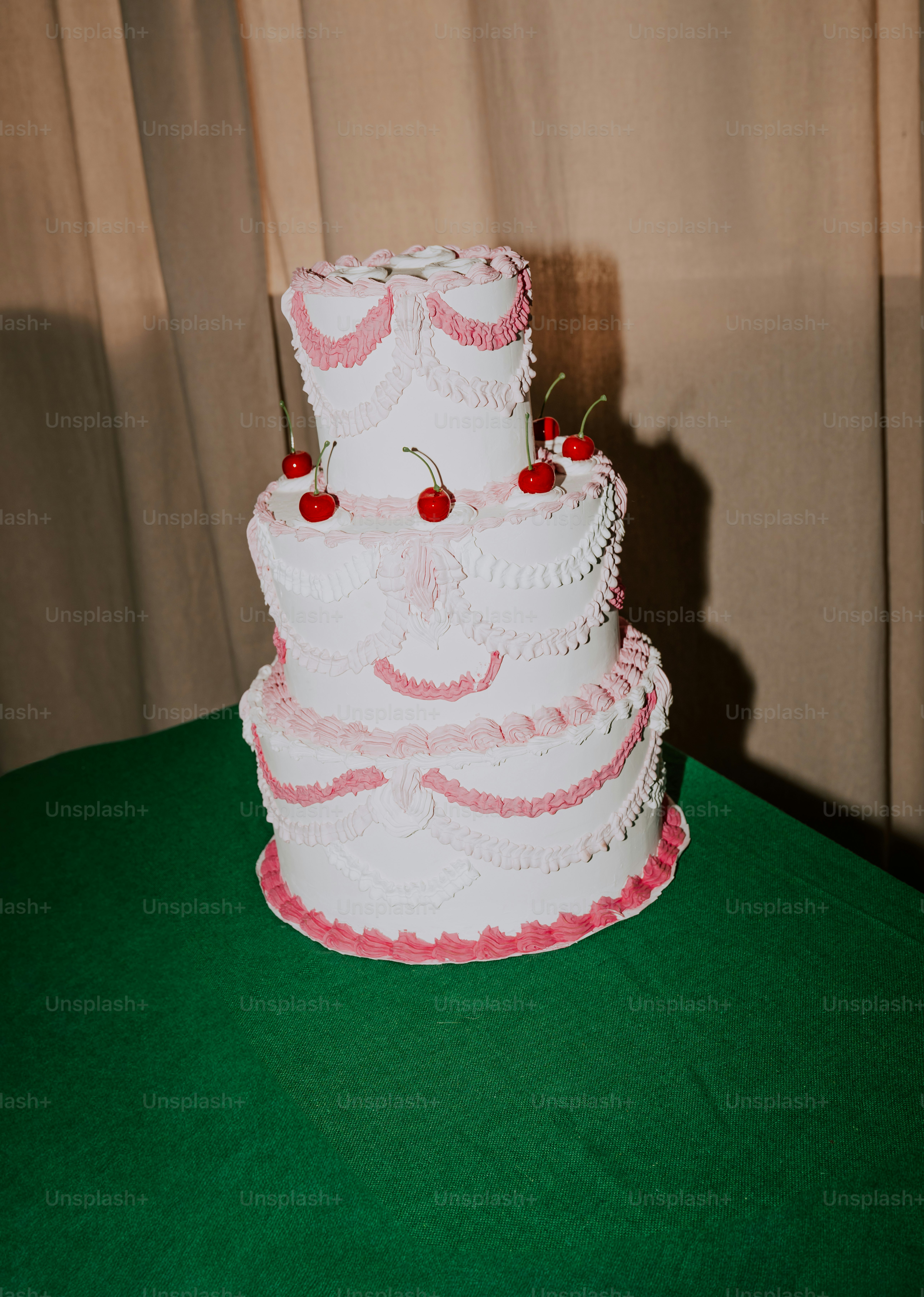 A three-tiered white cake decorated with pink frosting swags and red cherries on a green tablecloth
