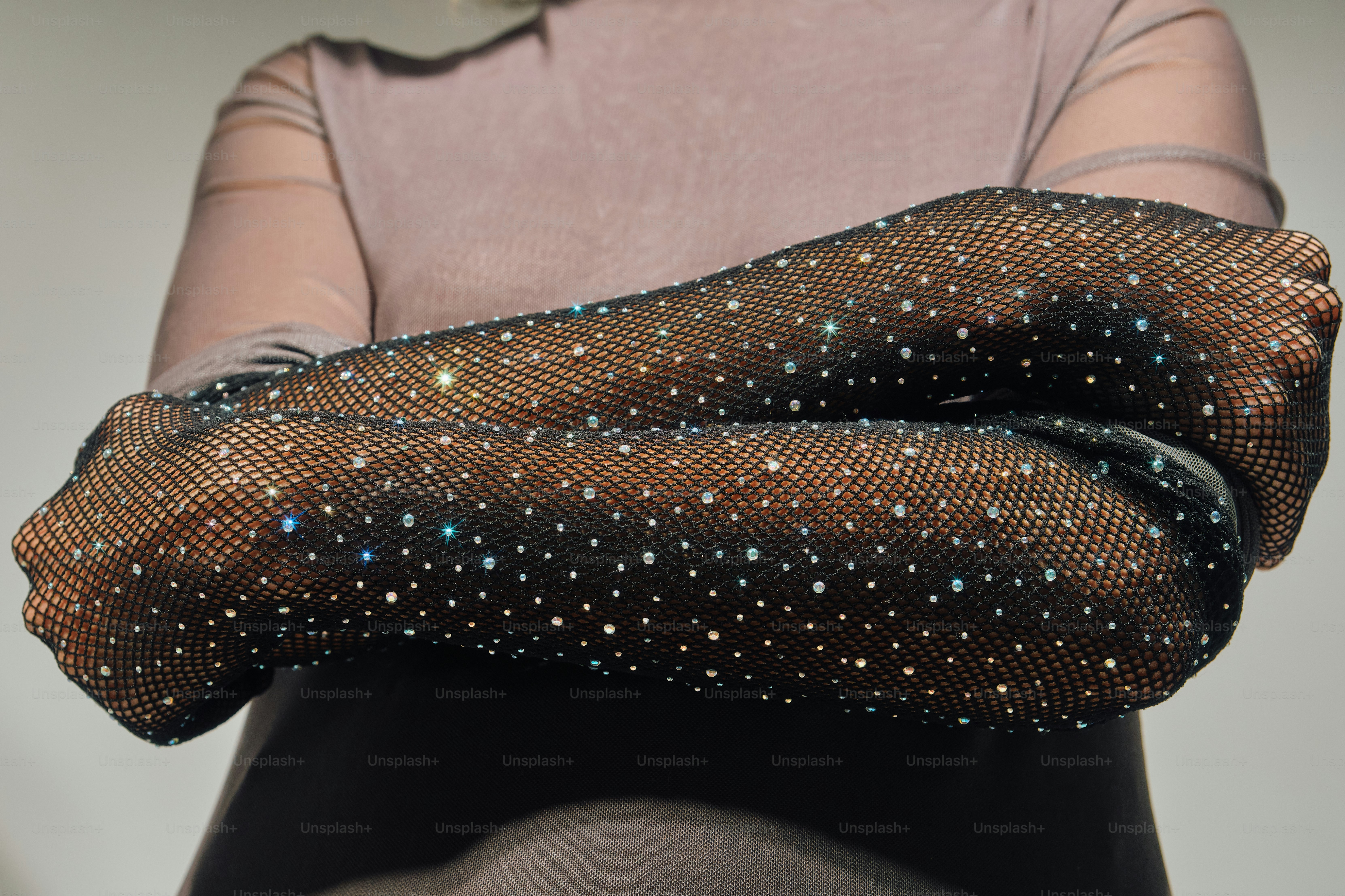A person wearing black fishnet gloves embellished with sparkling rhinestones against a neutral background
