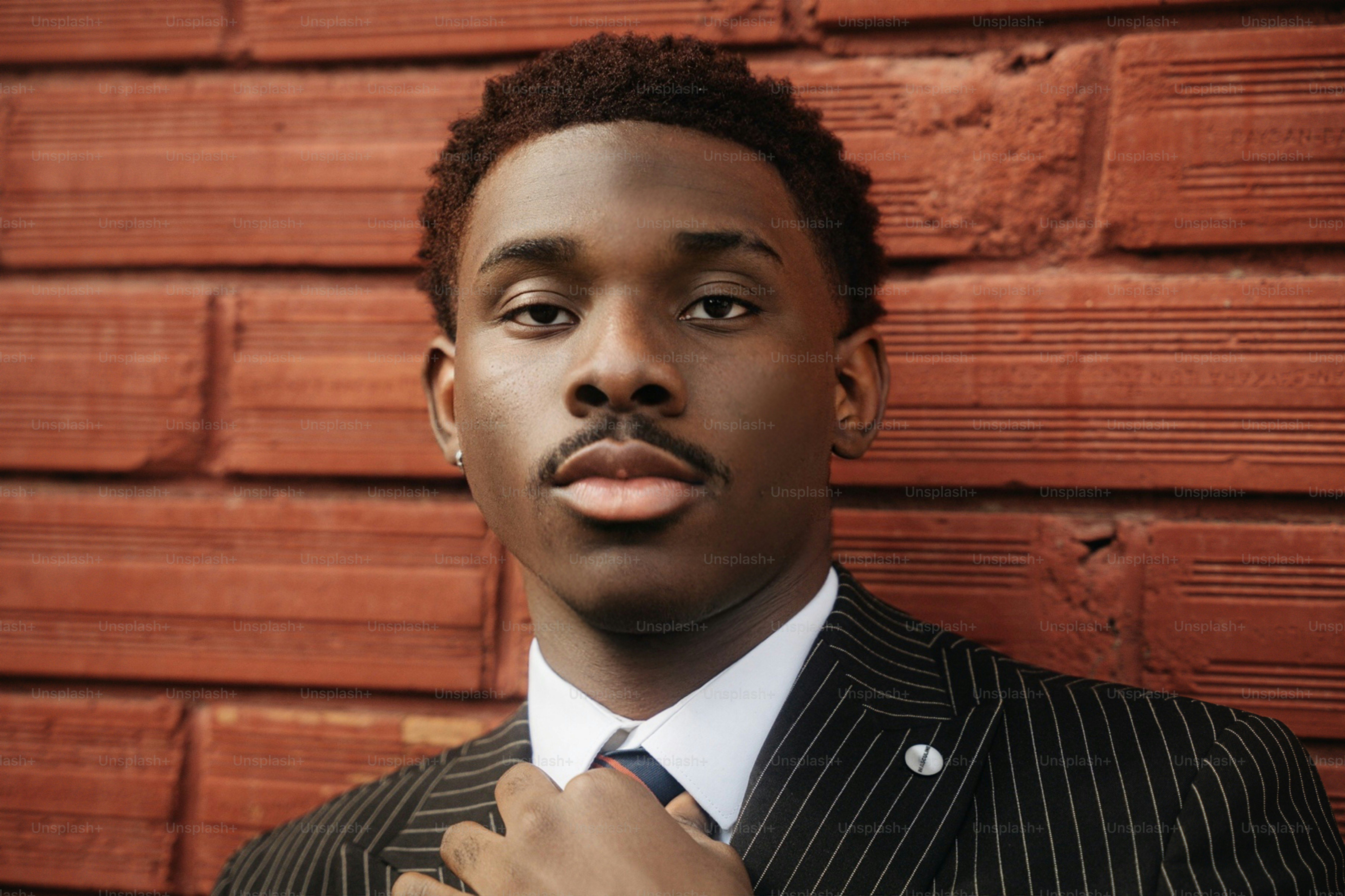 A young adult man in a pinstripe suit adjusting his tie against a red brick wall