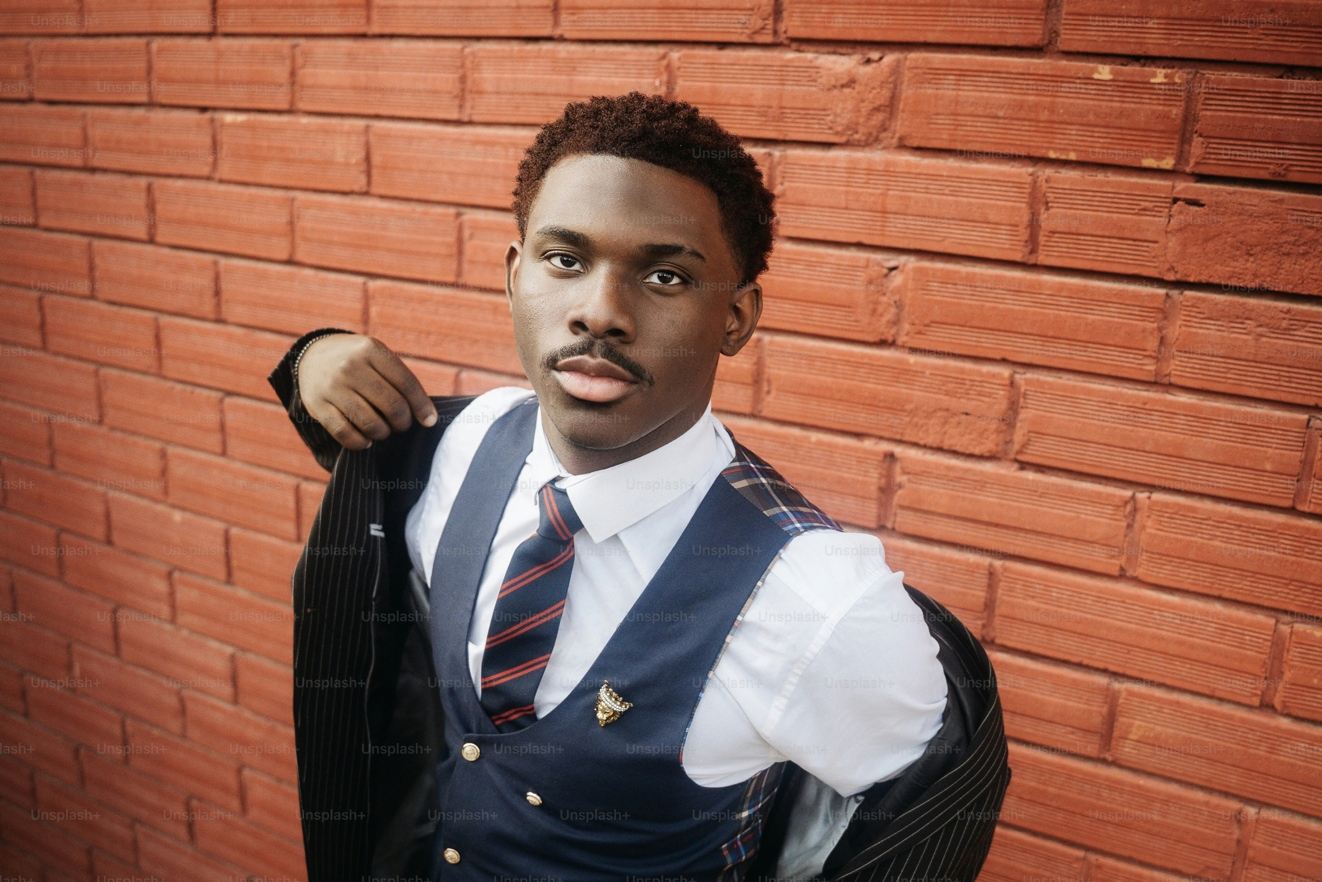 A young man in a white shirt, striped tie, and navy vest against a red brick wall