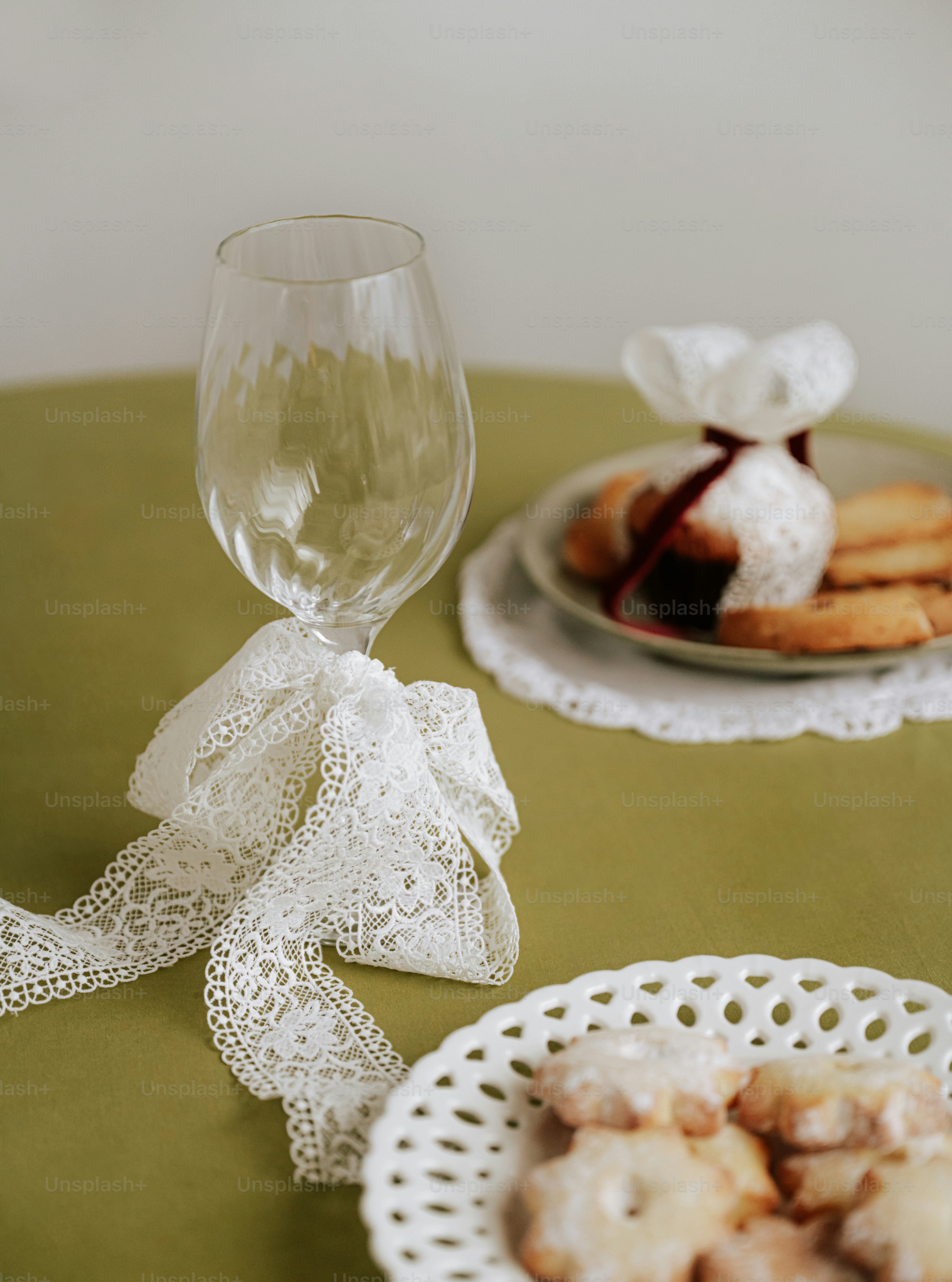 A wine glass tied with white lace ribbon beside plates of cookies on a green tablecloth