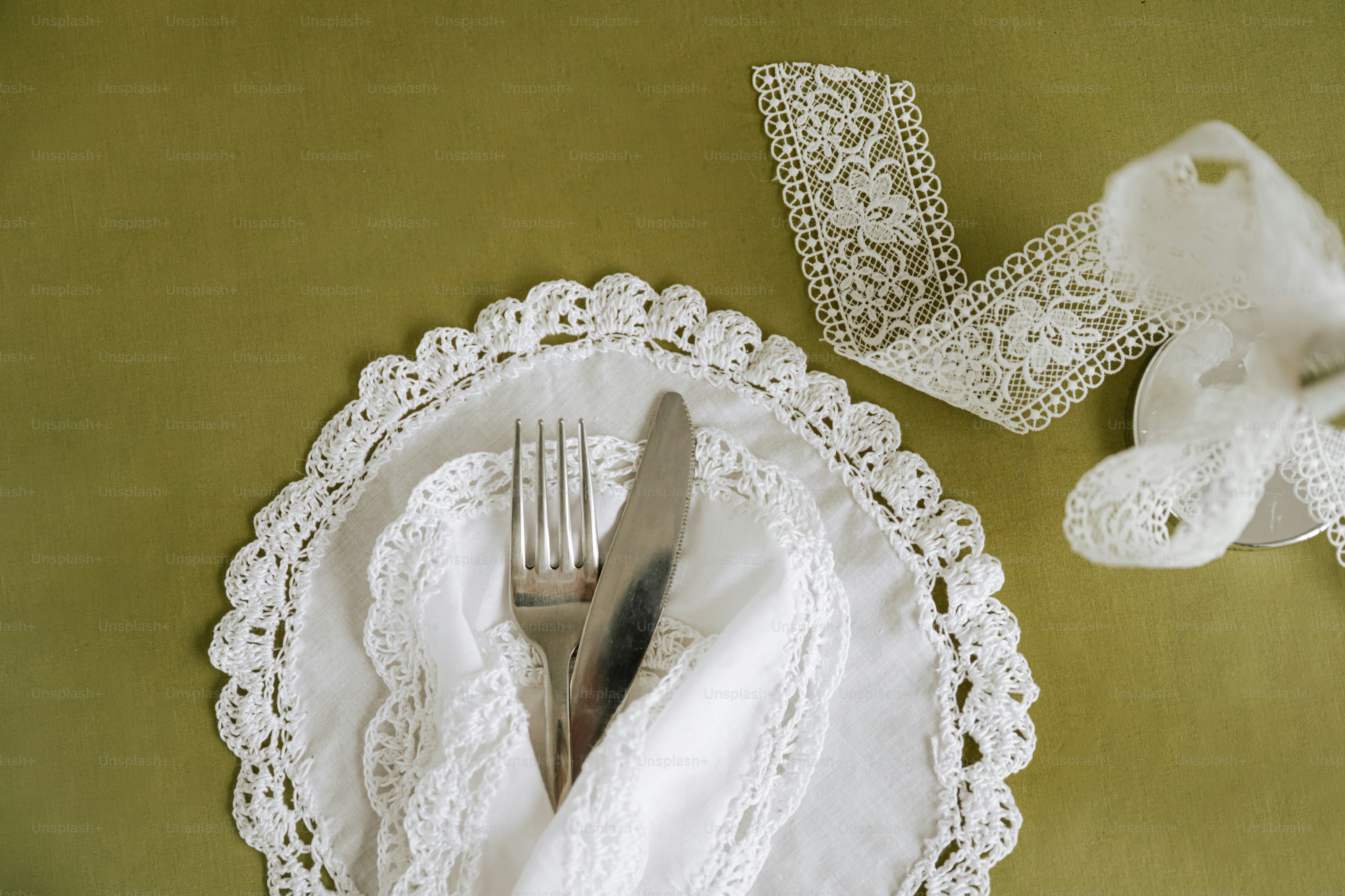 A silver fork and knife on a white lace doily over a green tablecloth