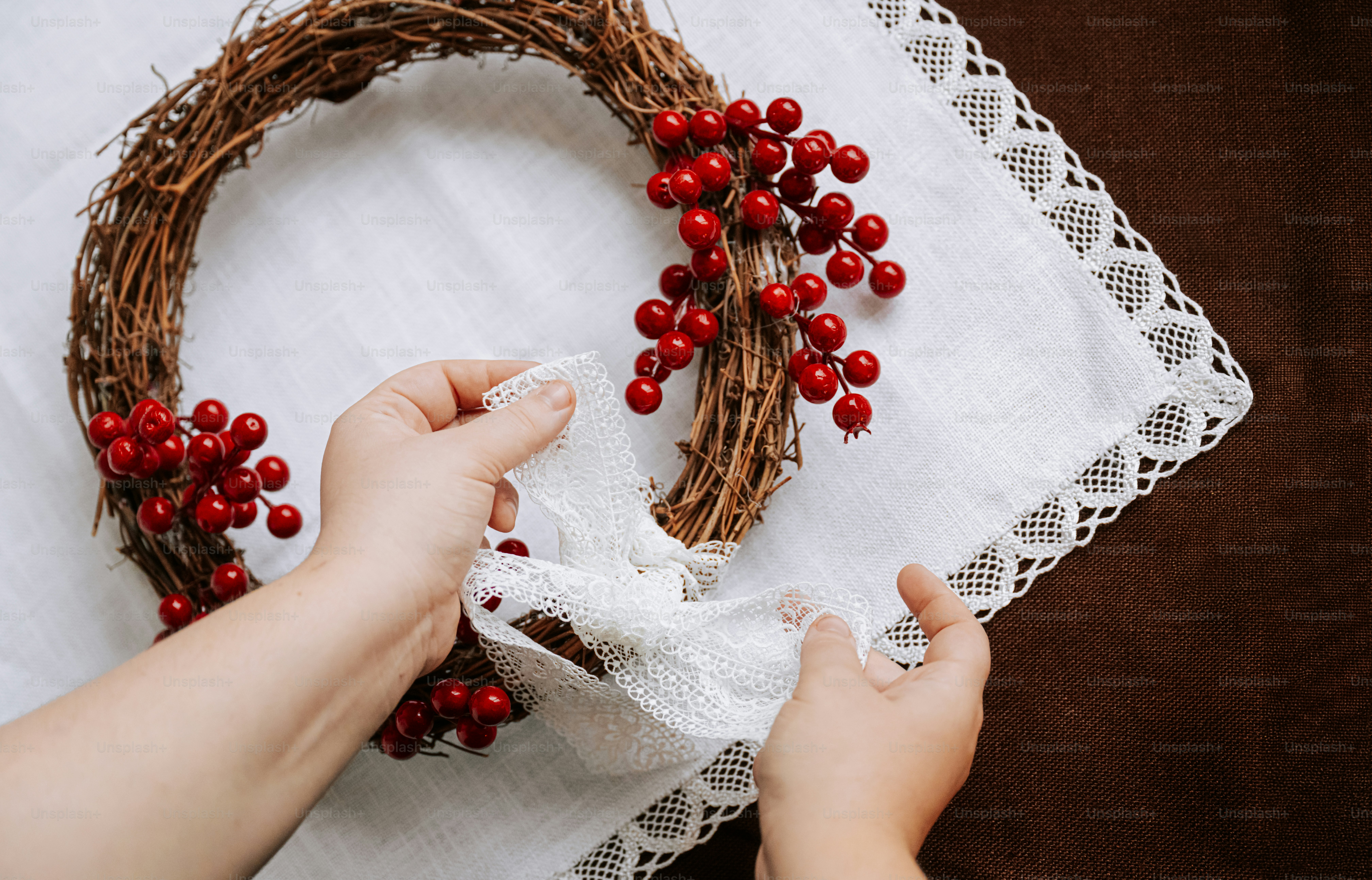 Hands tying a white lace ribbon onto a rustic grapevine wreath with red berries