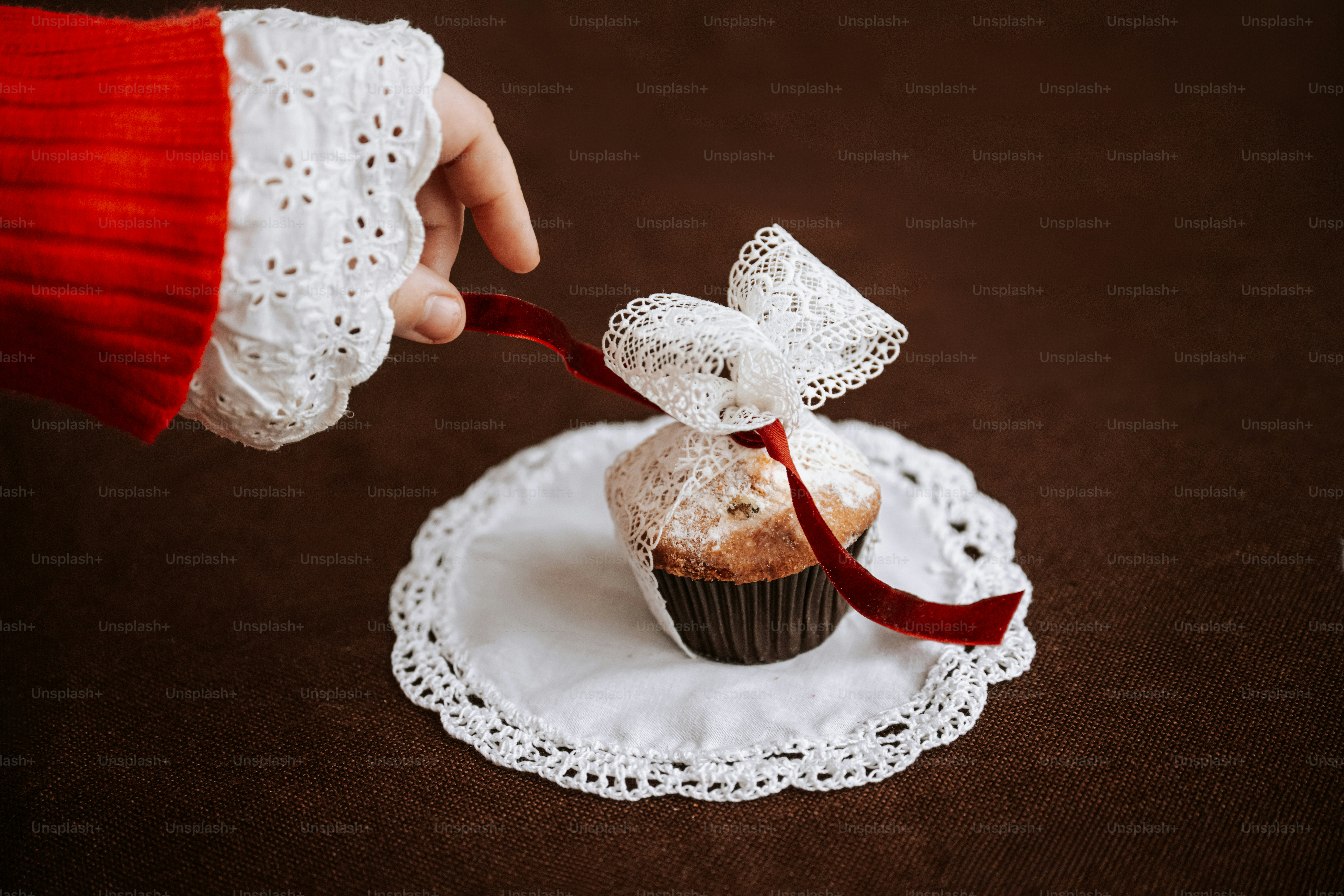 A hand in a red sweater sleeve pulling a red ribbon on a lace-wrapped muffin