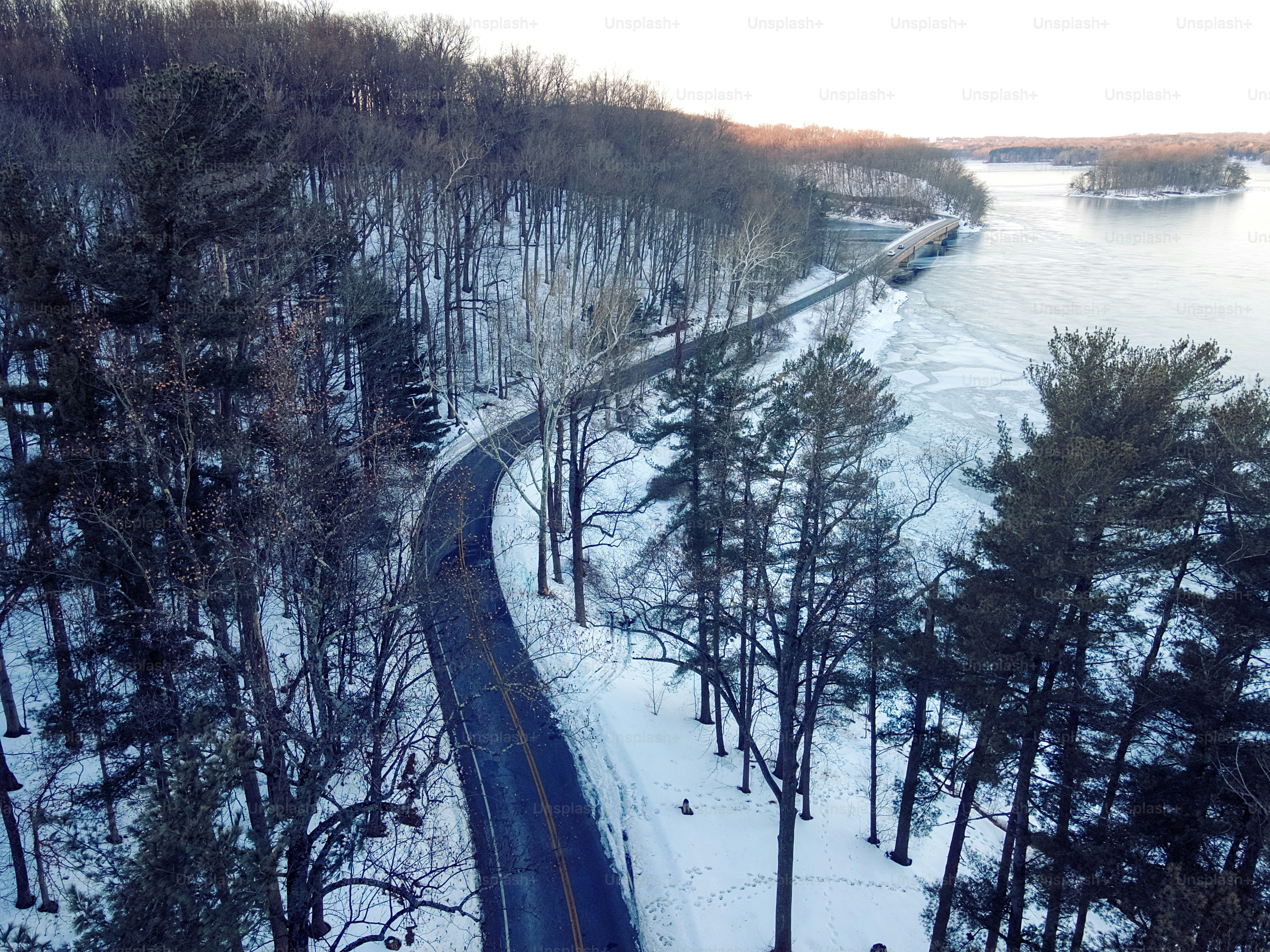 An aerial view of a winding road through a snow-covered forest beside a partially frozen lake