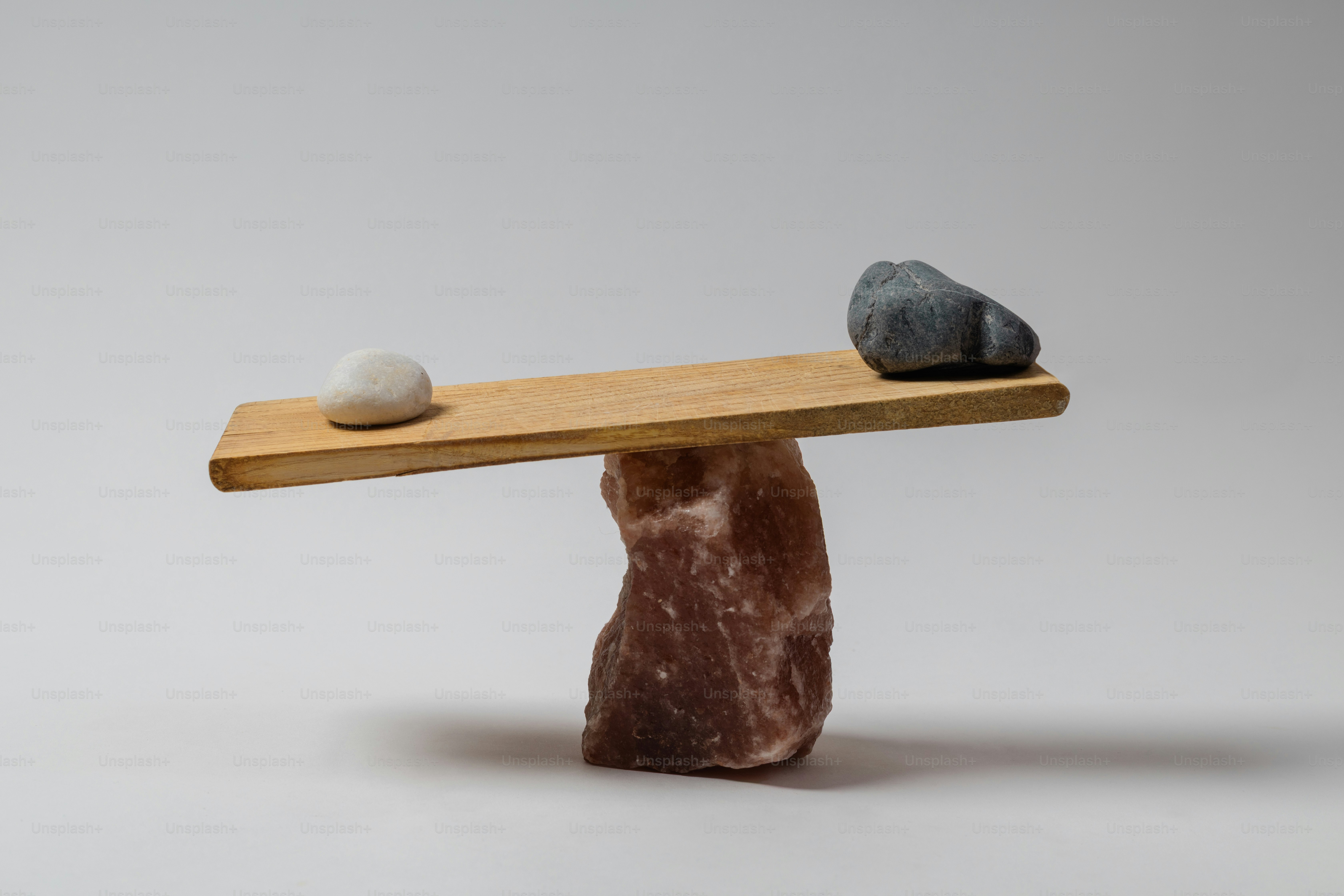 A wooden plank balanced on a large brown rock with a white stone and a dark stone