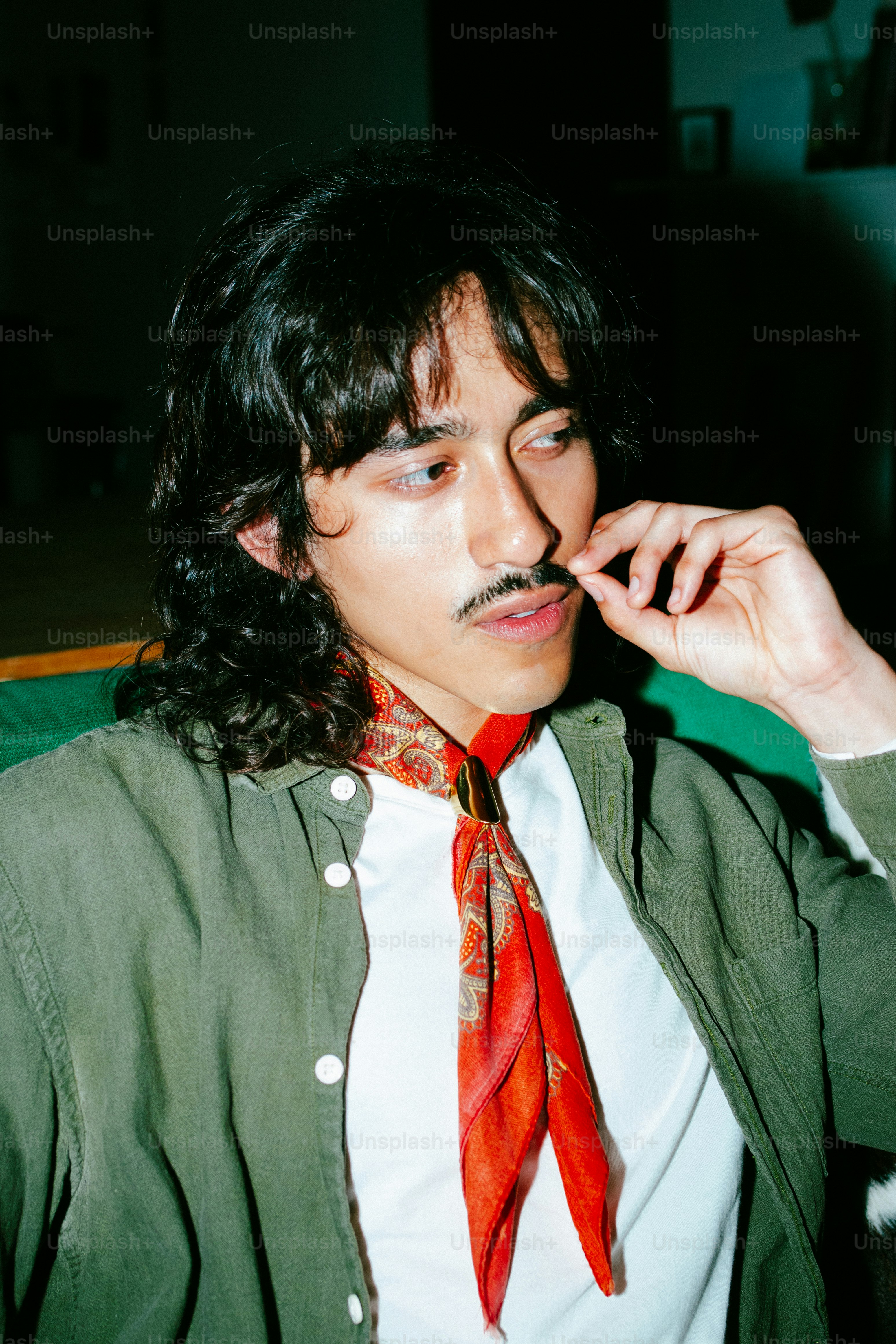 Un hombre de pelo largo y oscuro y bigote, con un pañuelo rojo paisley y camisa verde oliva