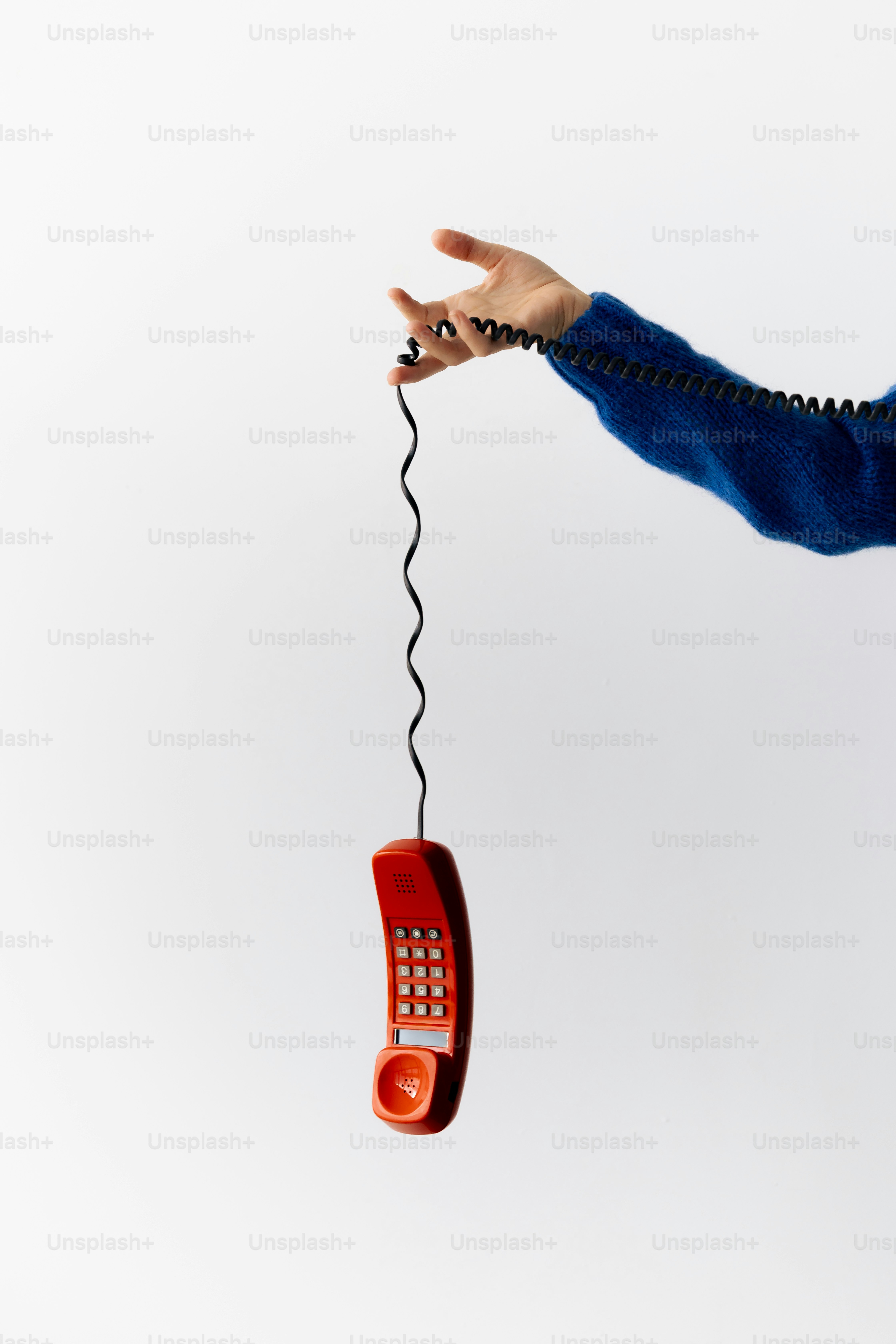 A hand in a blue sweater holding a red telephone handset by its coiled cord