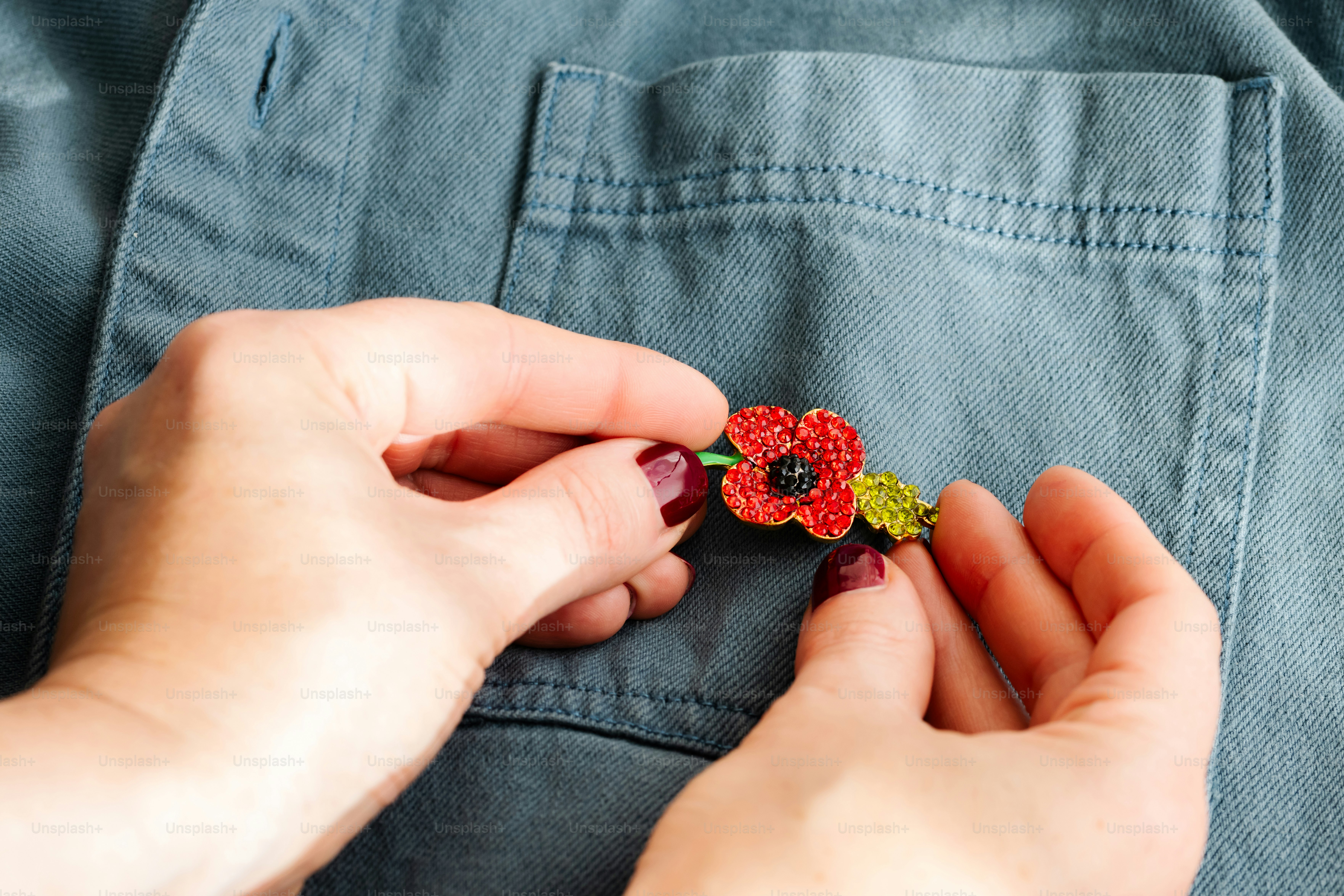 Hands pinning a red poppy brooch with green leaves onto a blue denim shirt