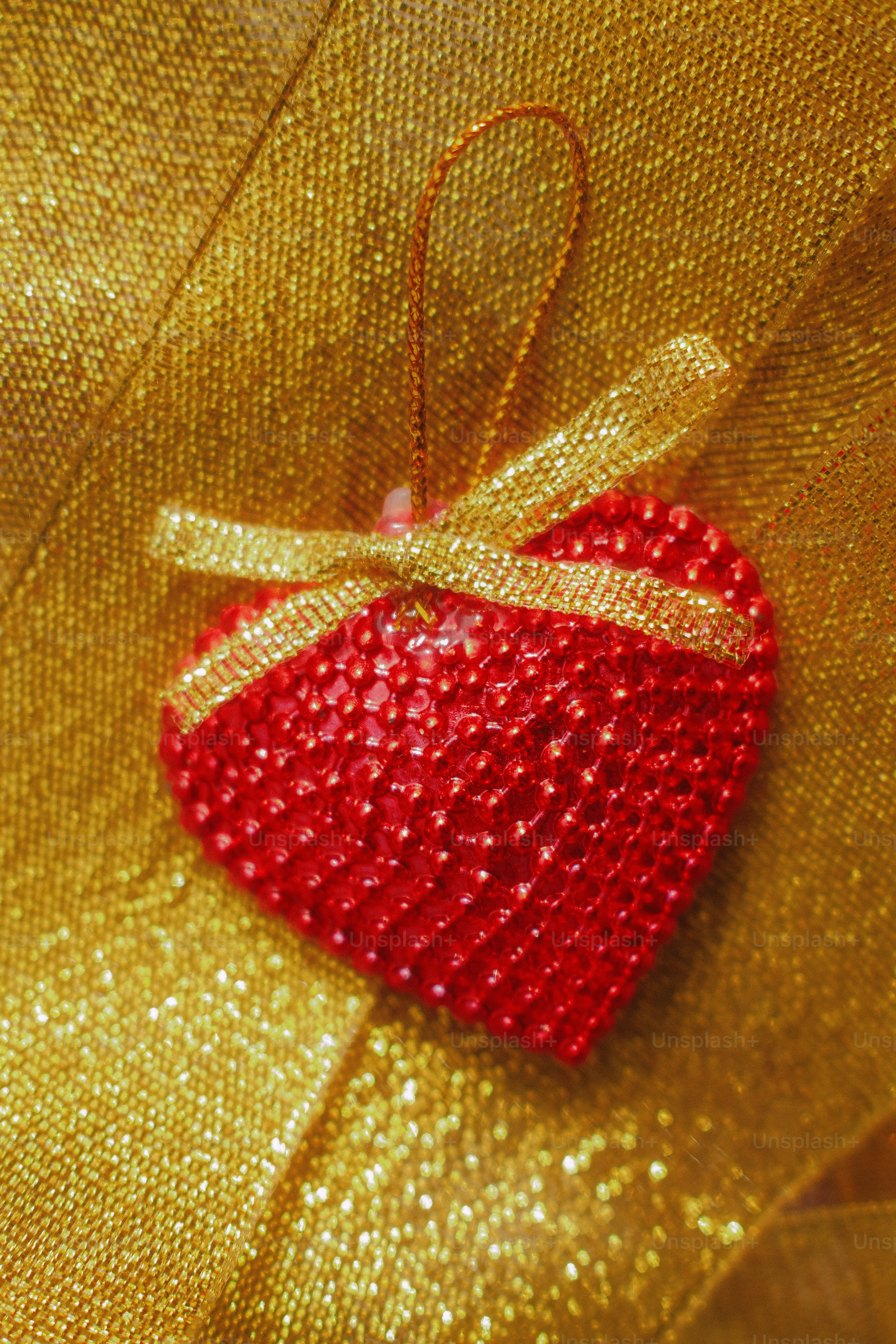 A red heart ornament covered in small beads with a gold ribbon against a shimmering gold background