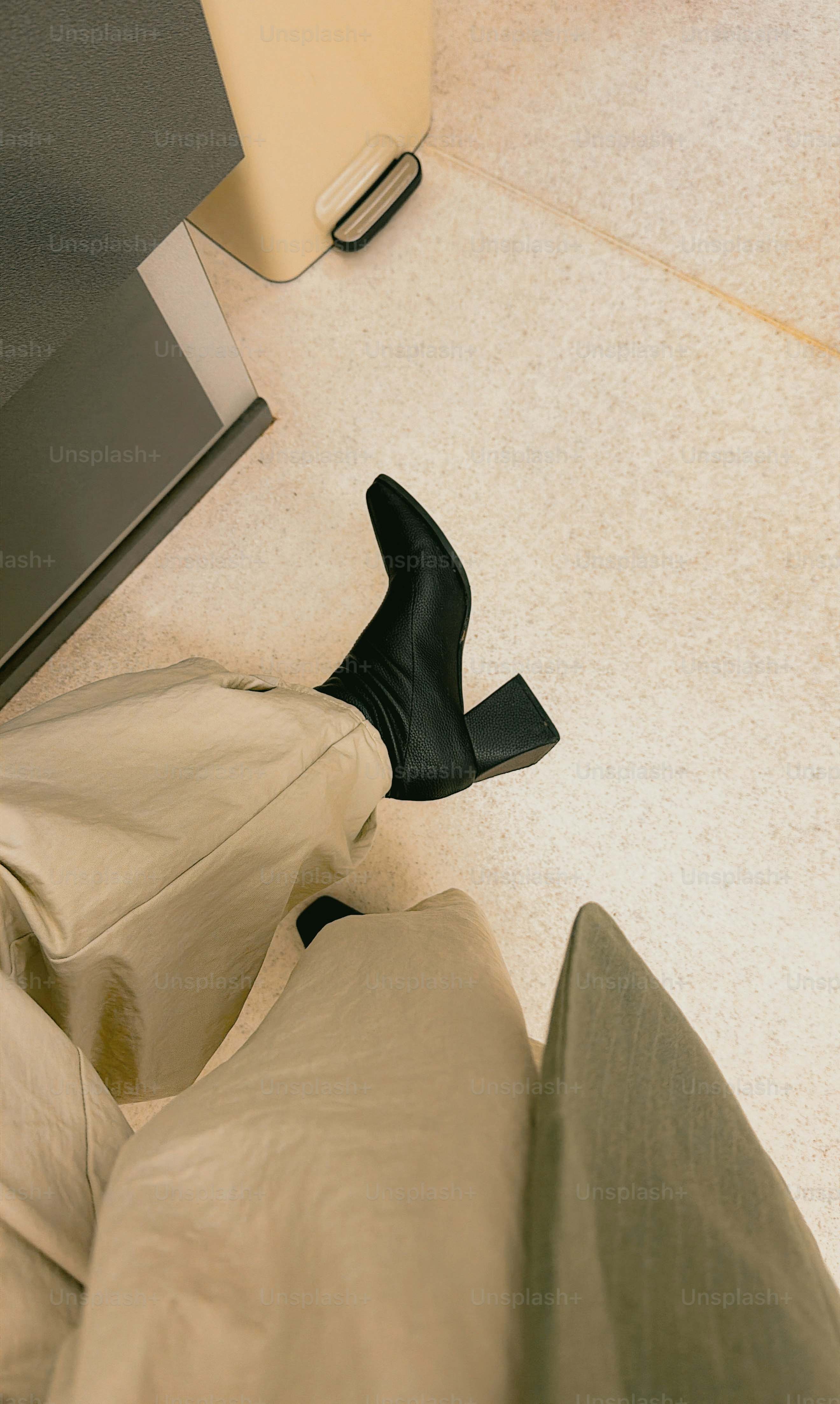 A person wearing beige trousers and black block-heeled boots on a light speckled floor