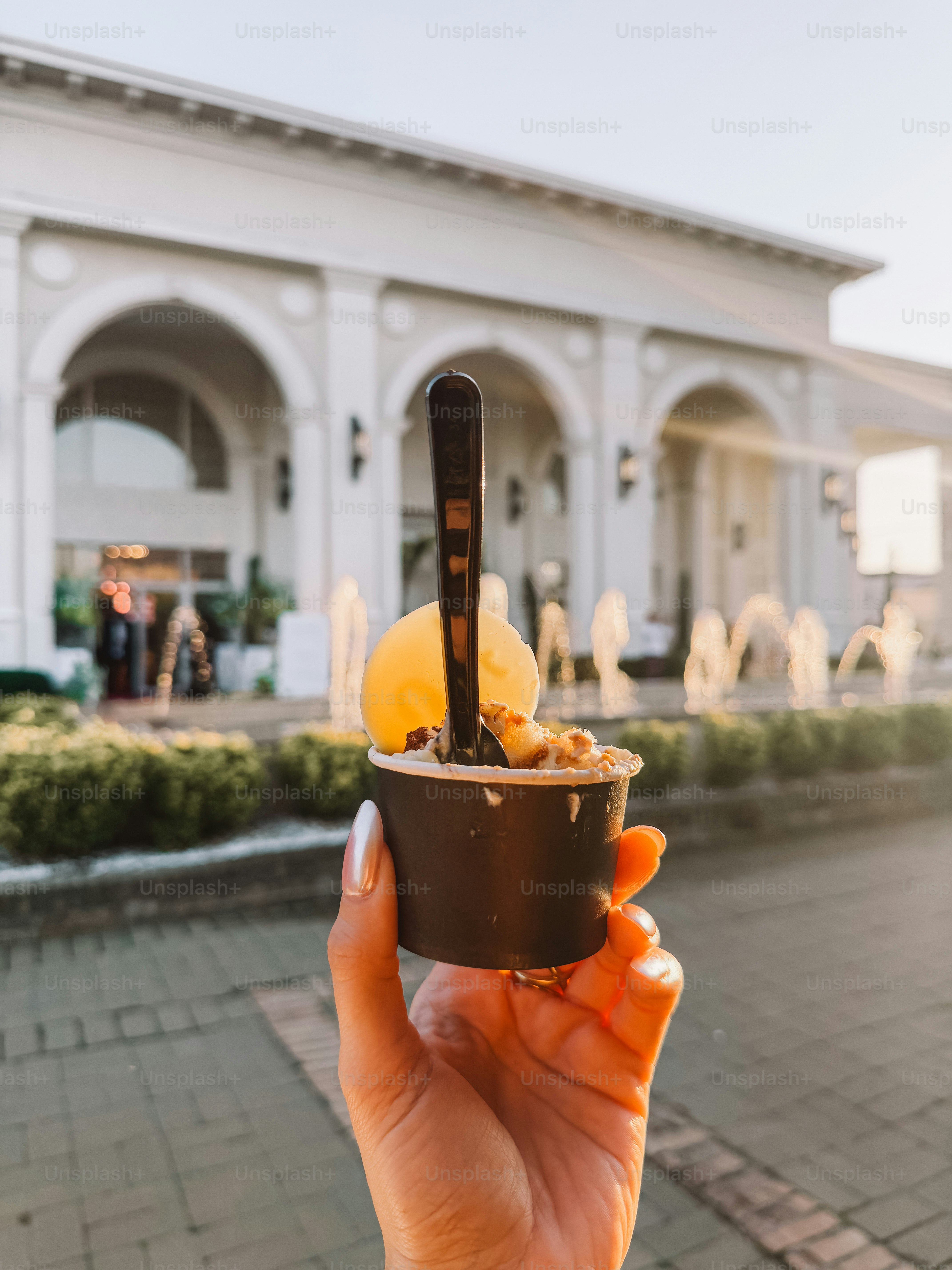 A hand holding a small cup of gelato with a spoon in front of a sunlit building