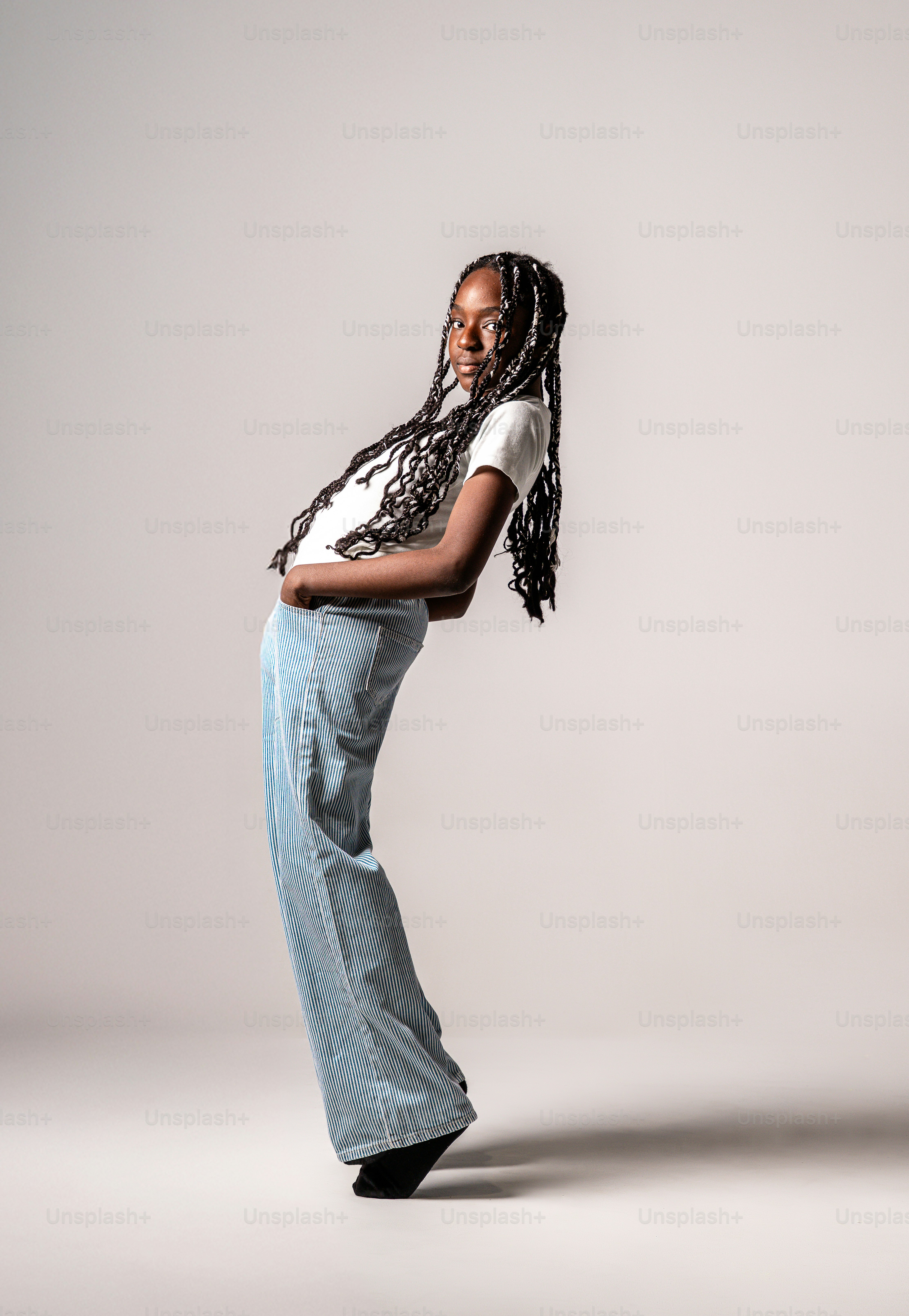 A teenager with long braids in a white t-shirt and striped trousers leaning back