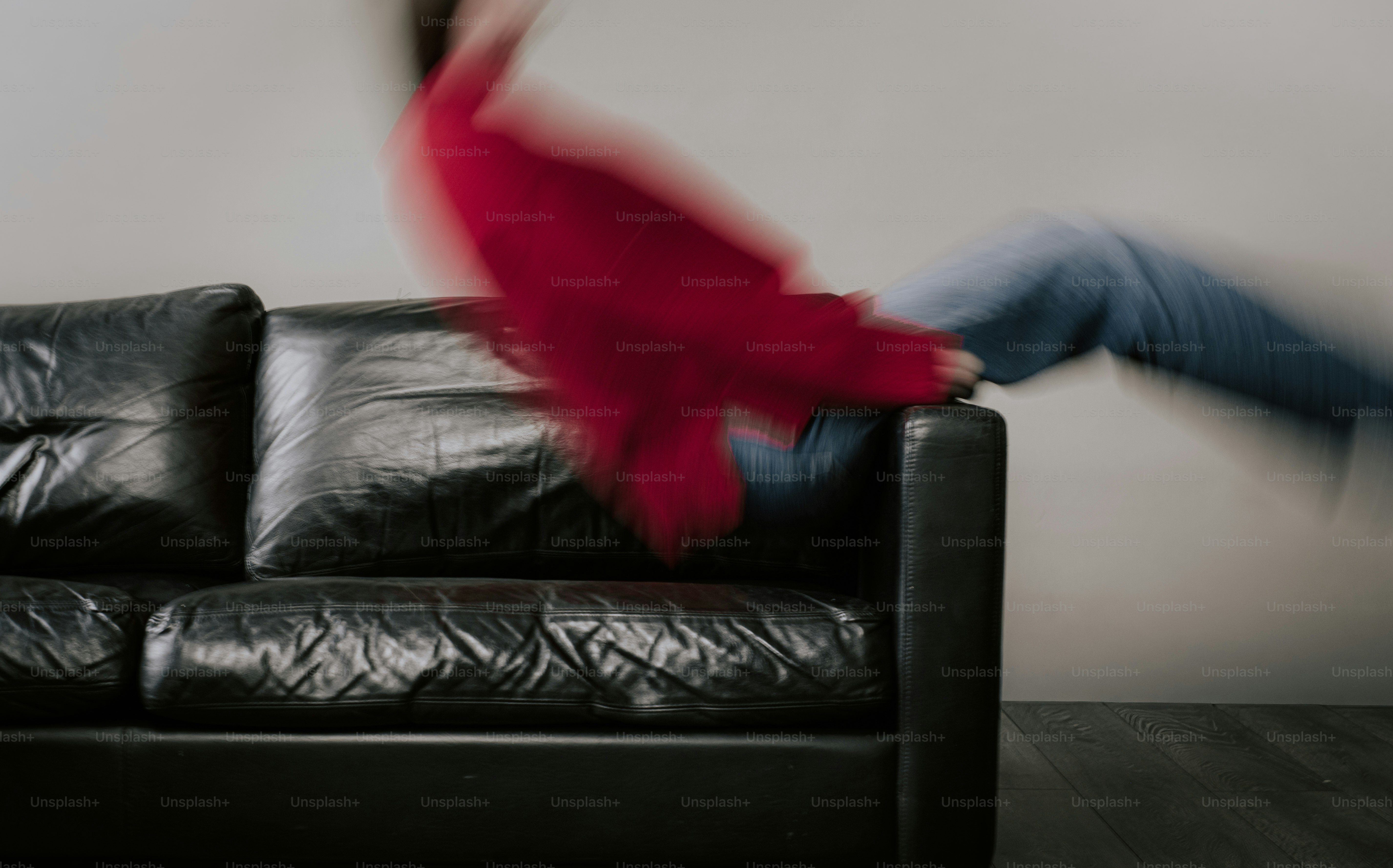 A person in a red sweater and blue jeans jumping onto a black leather sofa