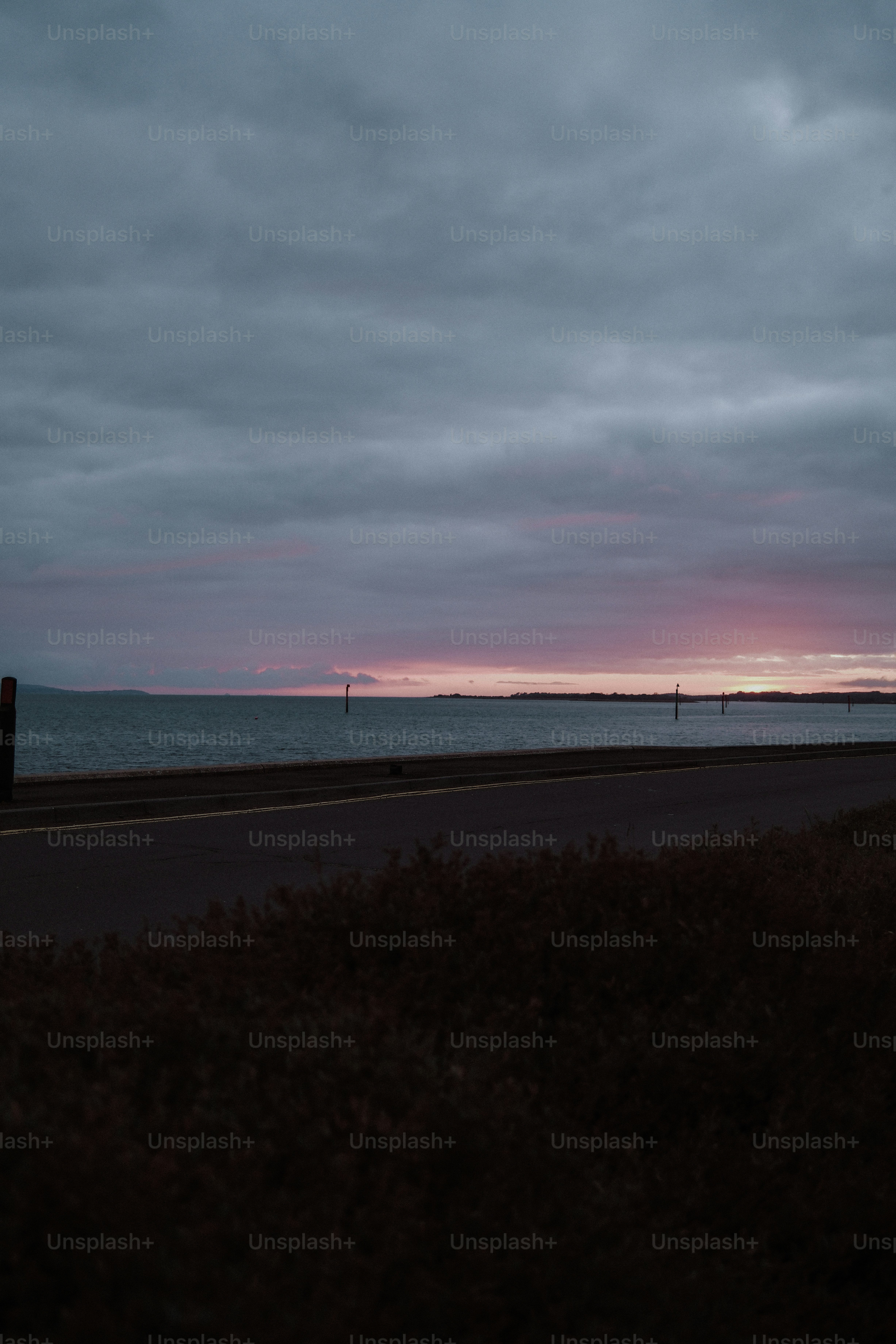 A dark road beside a calm sea under a cloudy sky with a soft pink glow